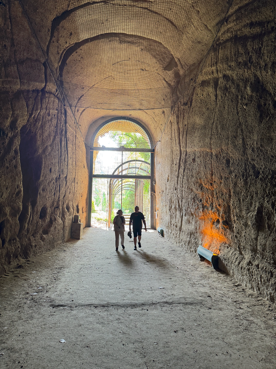 Two people walk through the tall, dimly lit Seiano Tunnel carved into rock, heading toward a bright opening framed by greenery and iron gates.