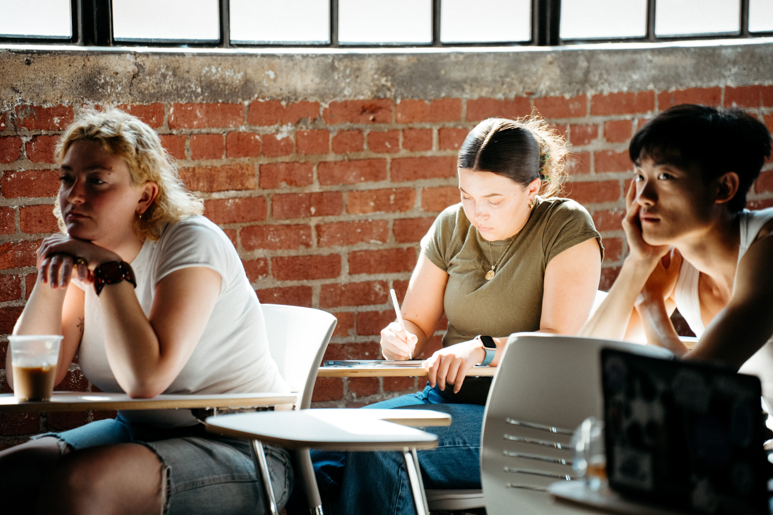 A student writes at a desk in class while two classmates beside them listen attentively.