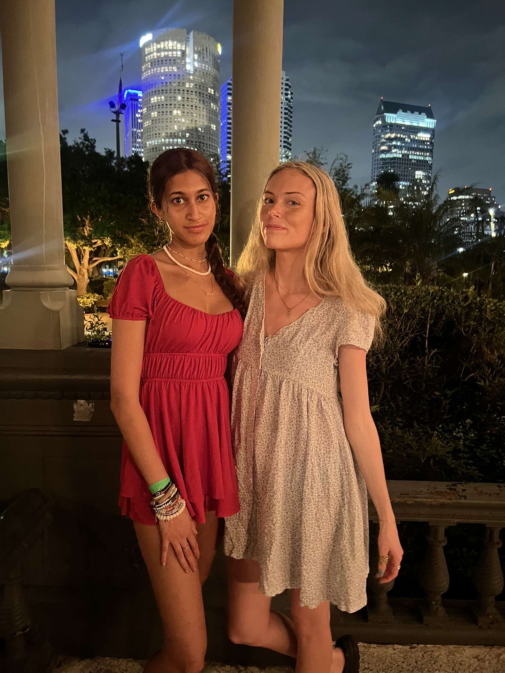 Two girls wearing dresses posed in front of a city skyline with palm trees in the background.