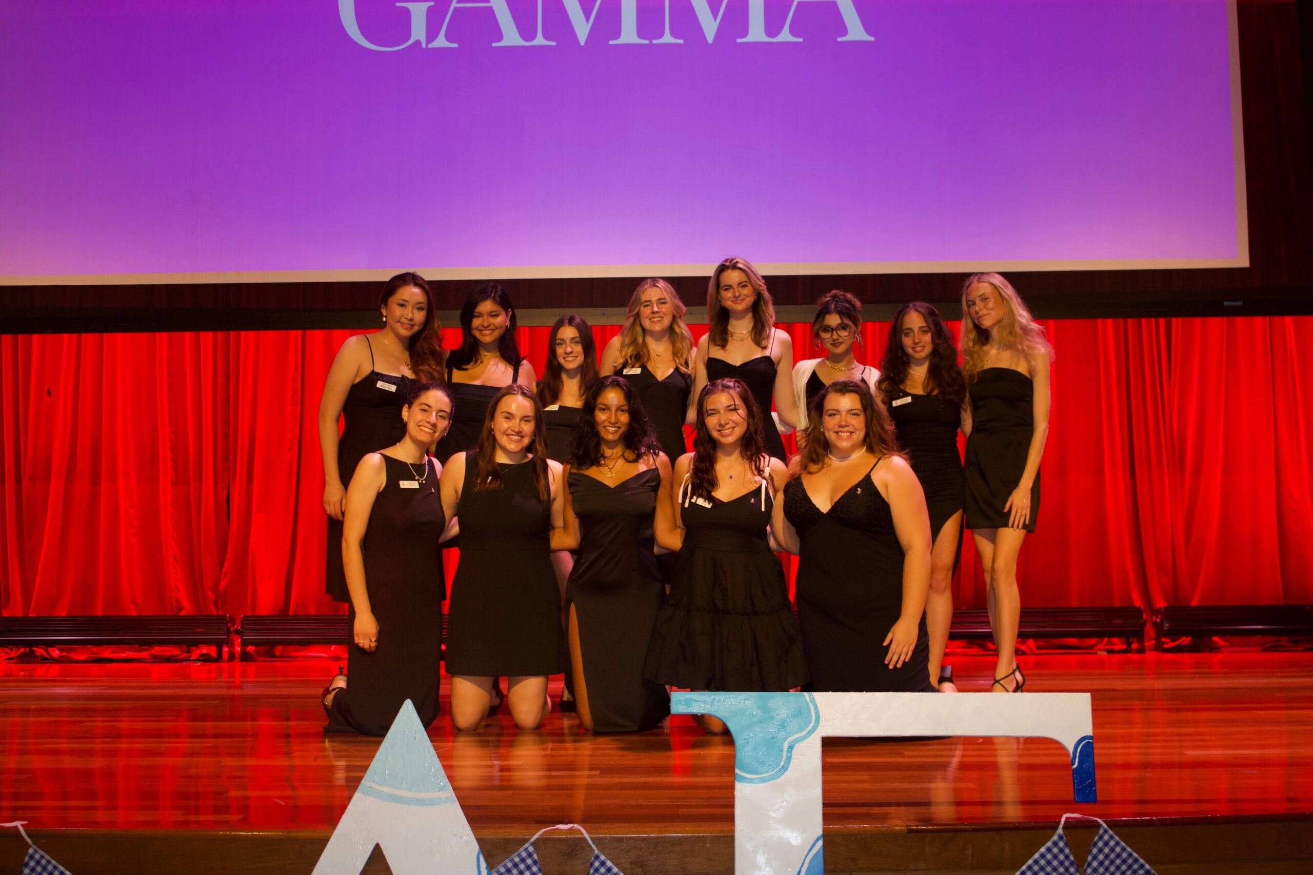A group of sorority sisters smiling in black dresses on a stage behind their letters.