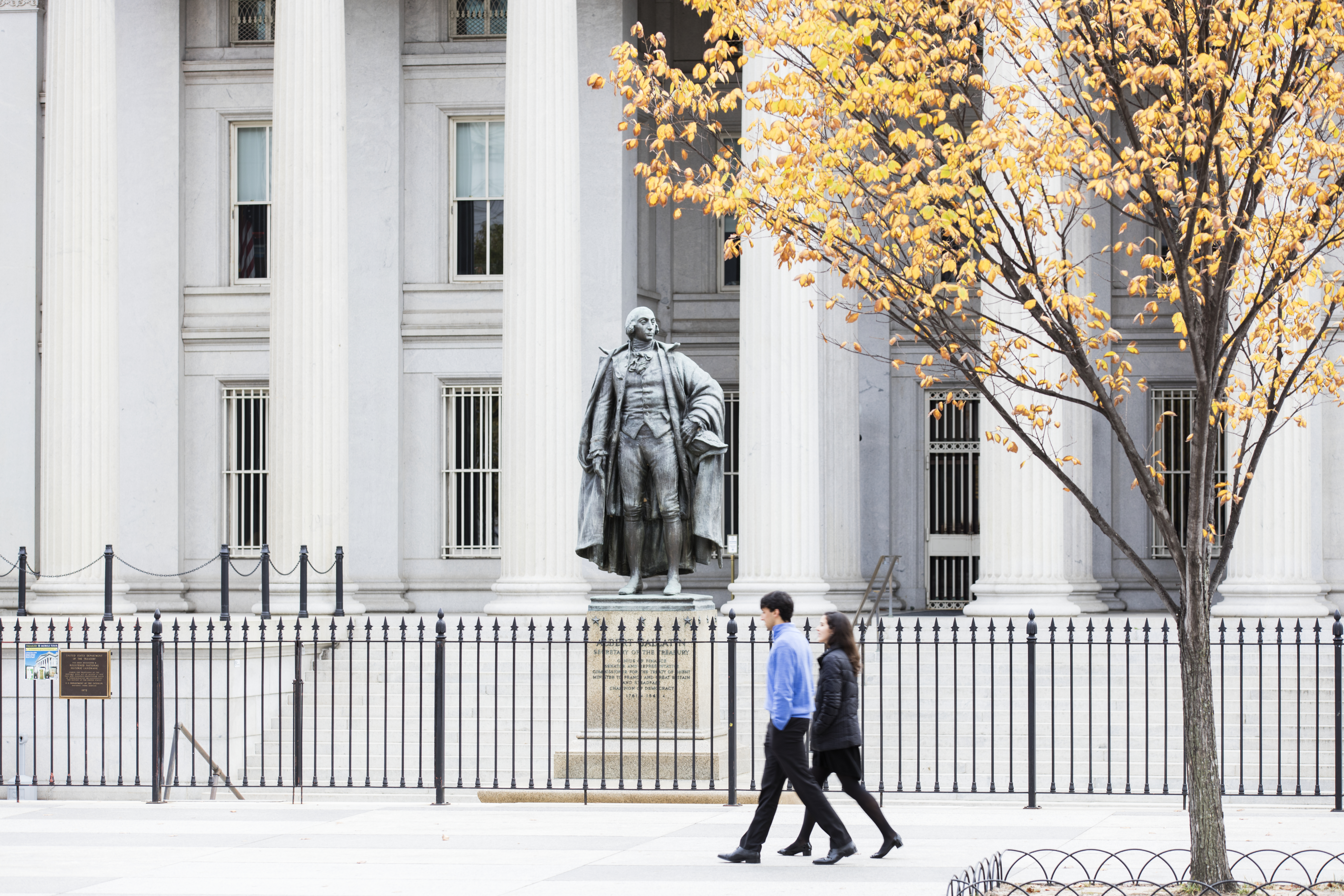 A statue of Alexander Hamilton stands in front of the US Treasury Building in Washington, DC, as two people walk past.