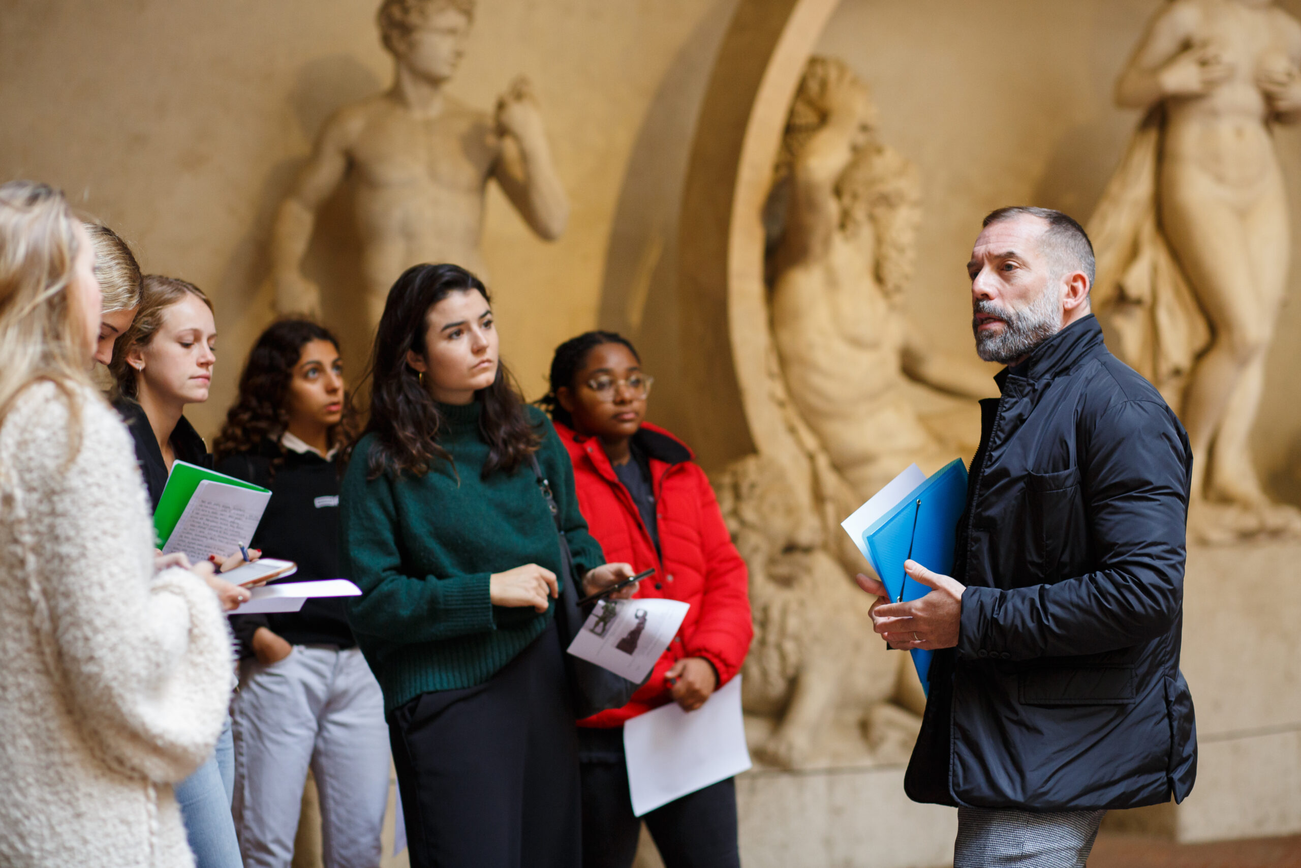 A group of students listen to an instructor while standing in front of large classical sculptures in a museum.