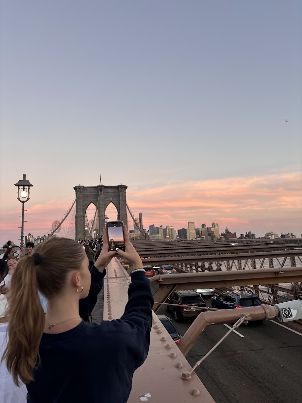 A girl facing away from the camera taking a photo of a pink & blue sunset over the Brooklyn Bridge