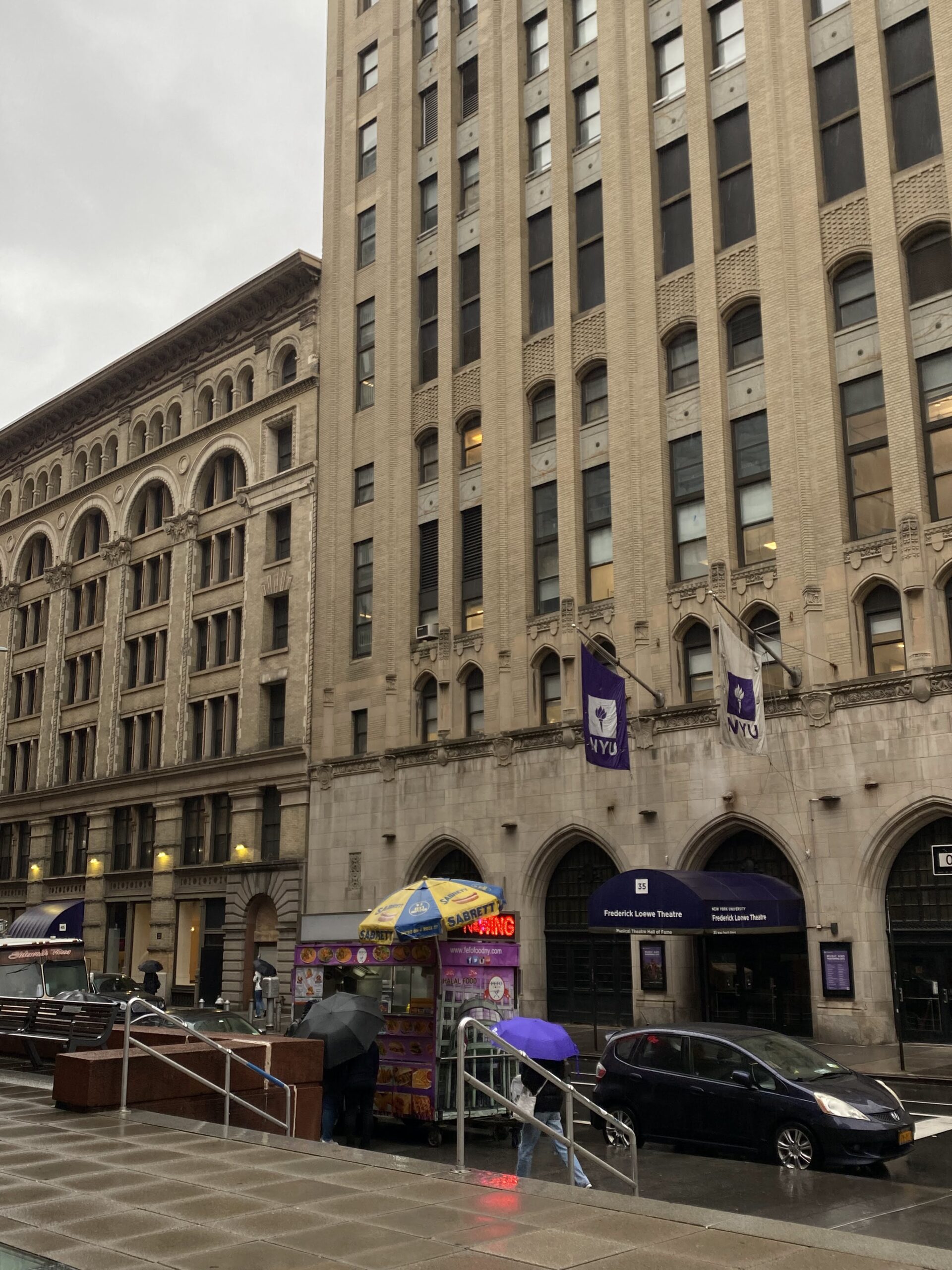 Photo of a street on NYU's campus showing tall buildings with NYU flags and a busy street below.