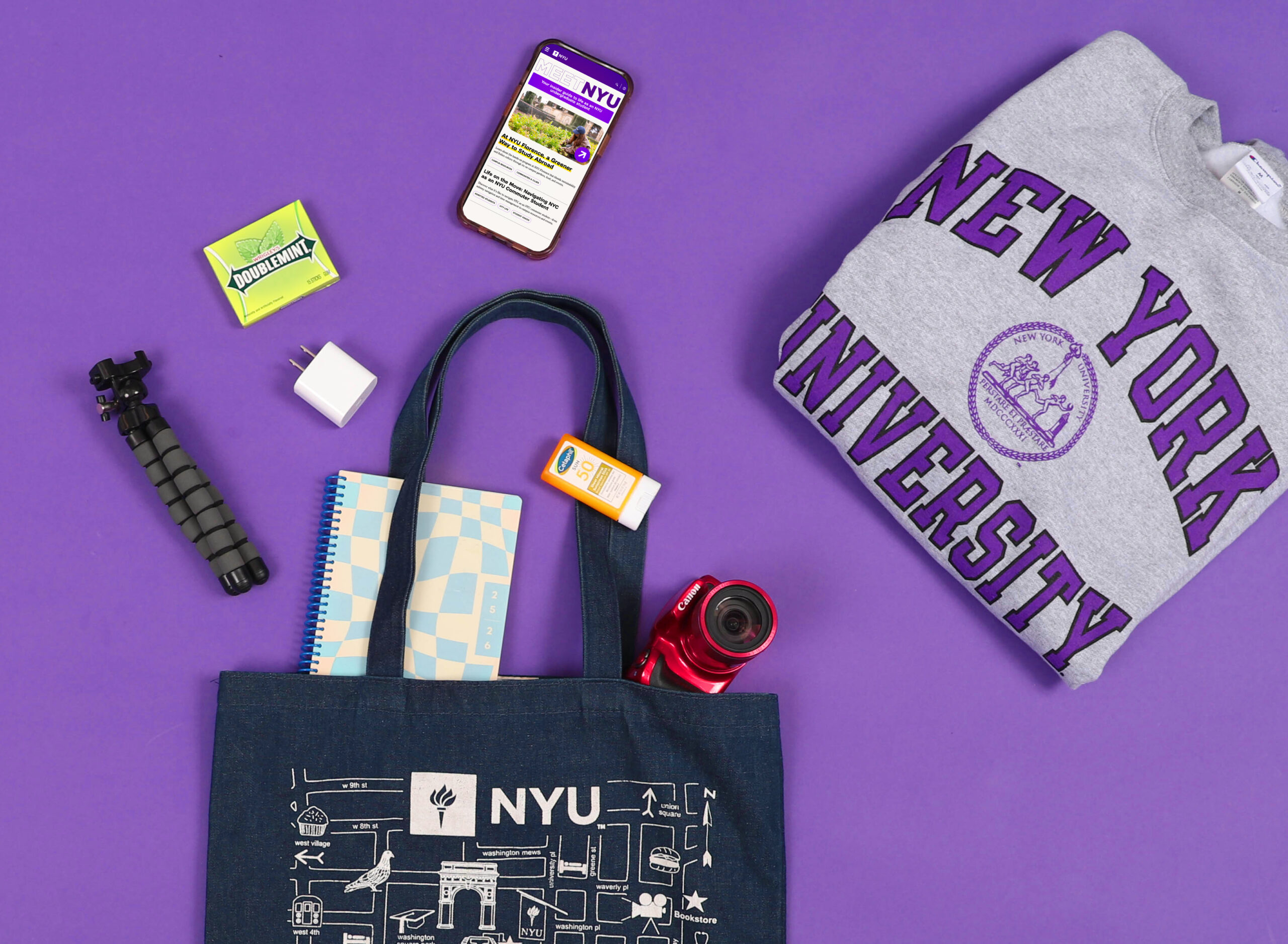 Flat lay of NYU-branded items: including a tote bag, notebook, phone, headphones, and stickers arranged on a purple background.