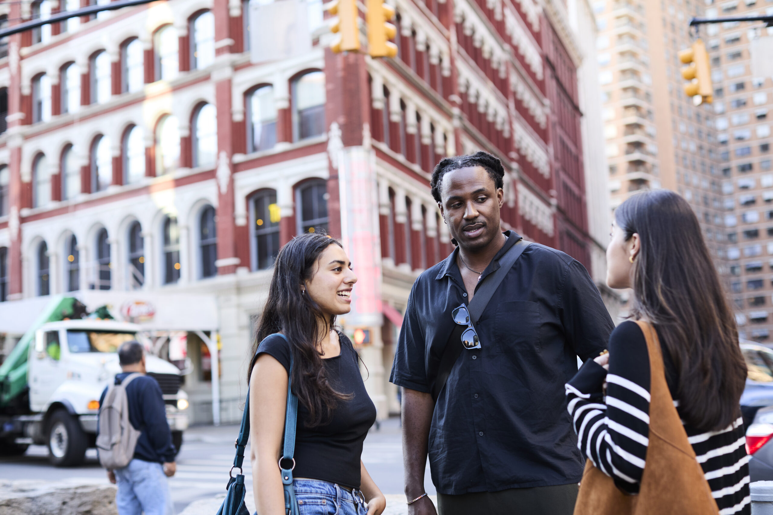 Three students standing on a busy NYC street, people and cars passing behind them.