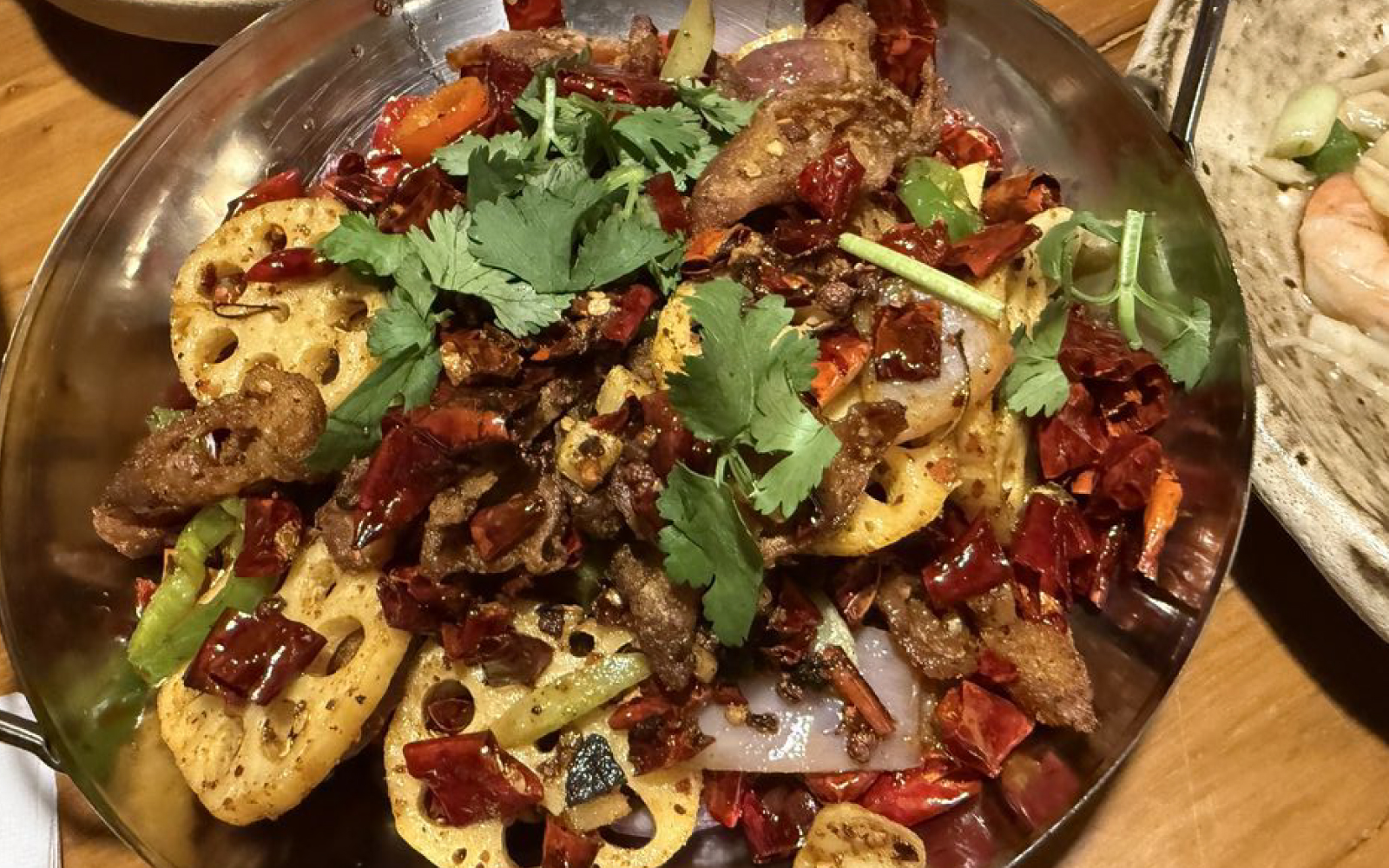 A metal bowl of spicy stir-fry with sliced meat, lotus root, dried red chilies, and cilantro on a wooden table.