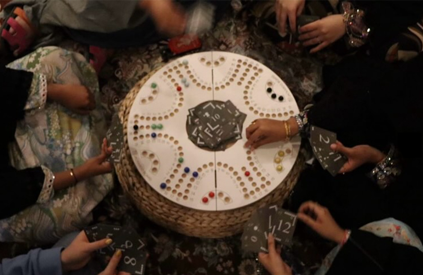 An overhead view of several people playing a round board game, with hands holding cards around a circular board.