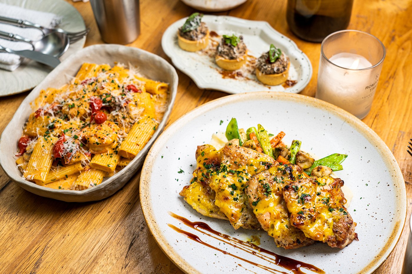 A meal featuring sauced grilled meat with vegetables, creamy rigatoni pasta, and small appetizers on a wooden table.