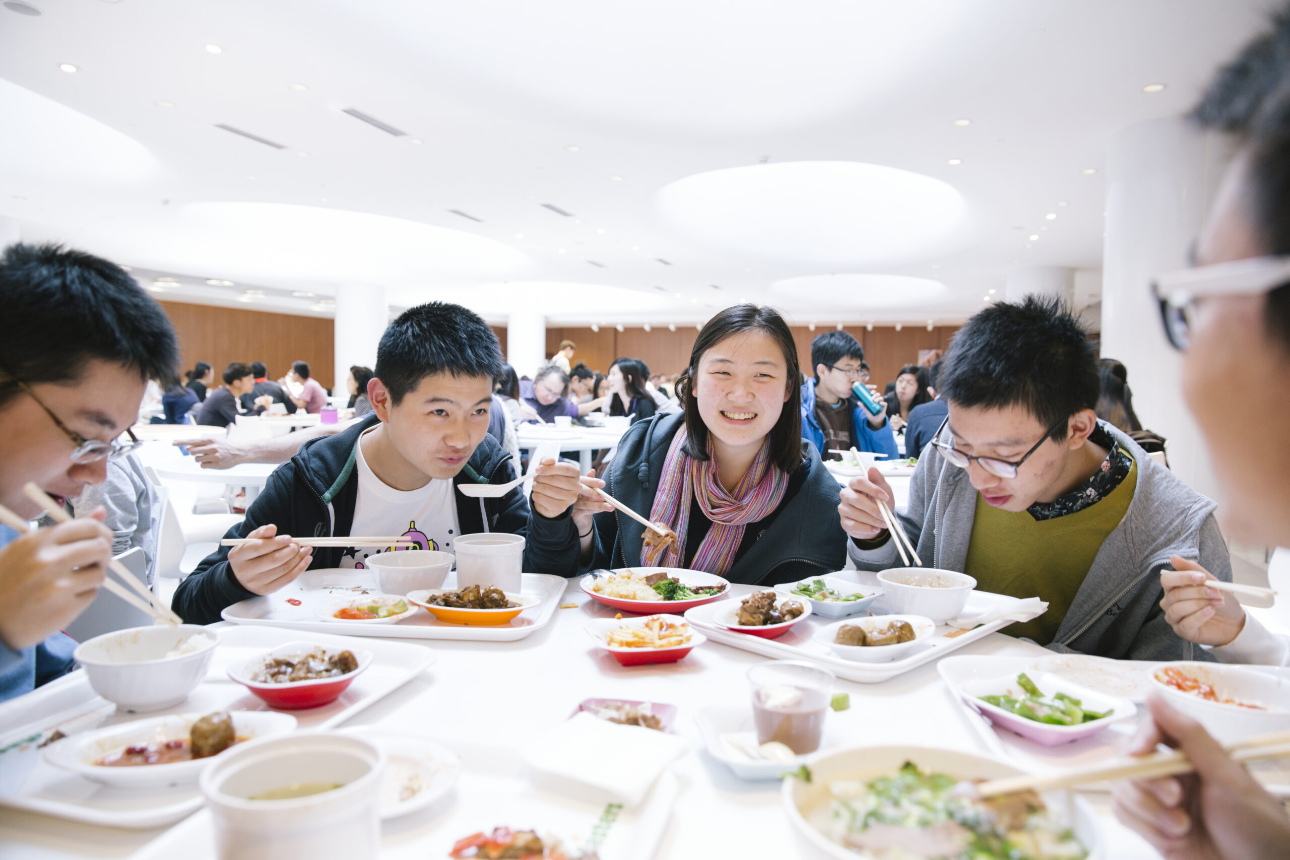 Students sitting together in a cafeteria, eating a meal and talking at a table filled with trays of food.