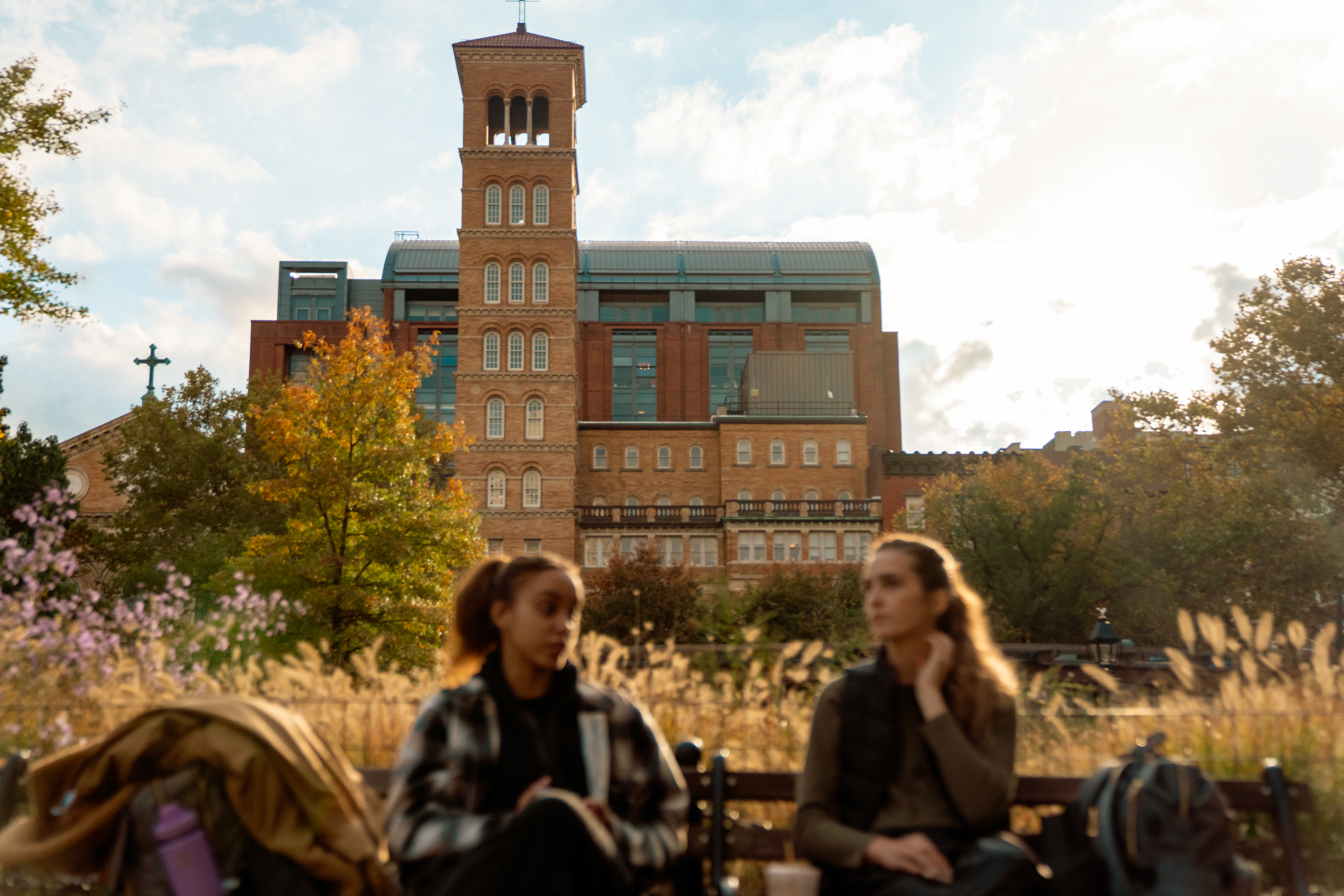 Two people sit on a bench in the foreground, slightly out of focus, with backpacks beside them and an NYU campus building in the background.