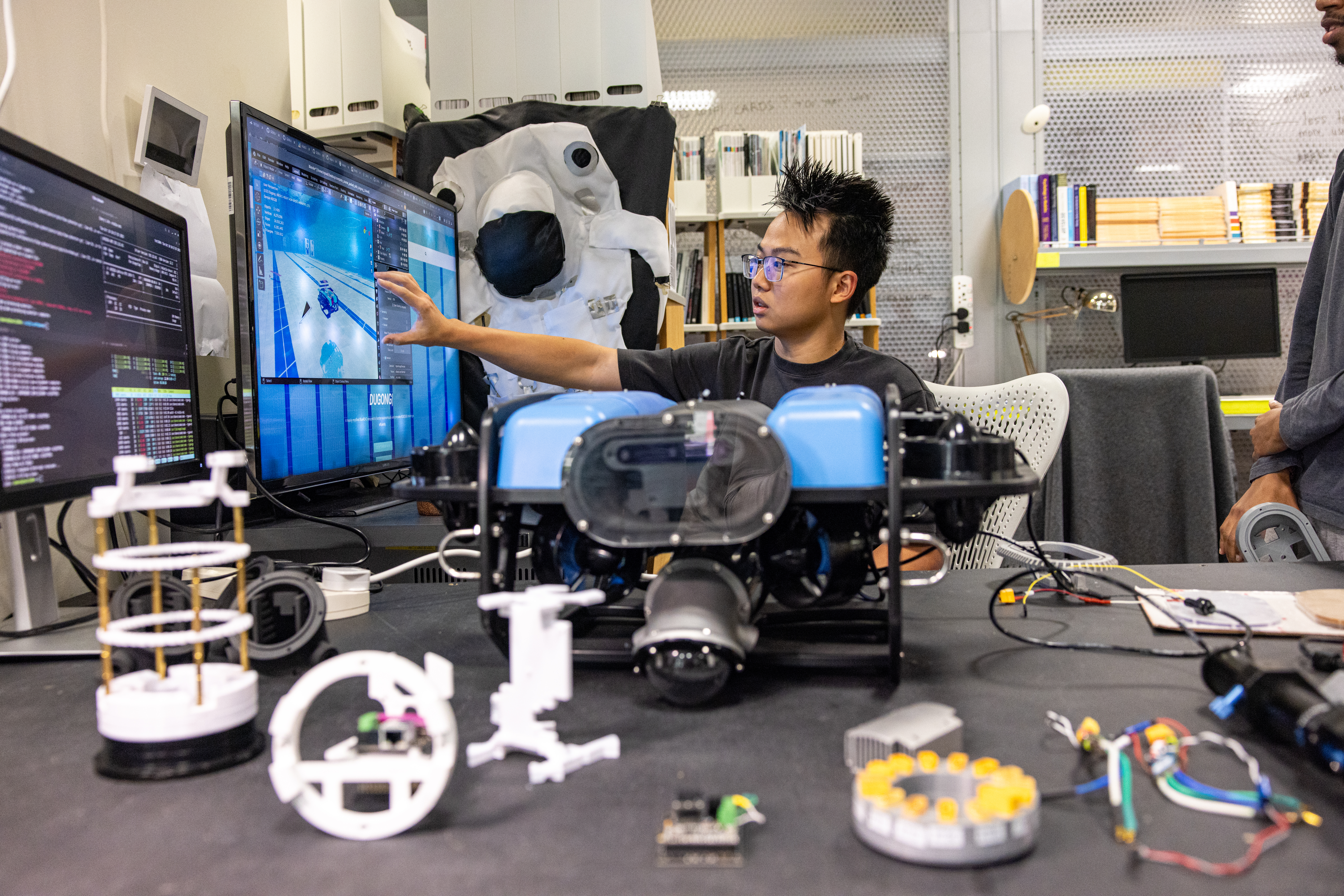 Students work on robotics in a lab, one pointing at code and simulations on monitors with a submersible robot and components on the desk.