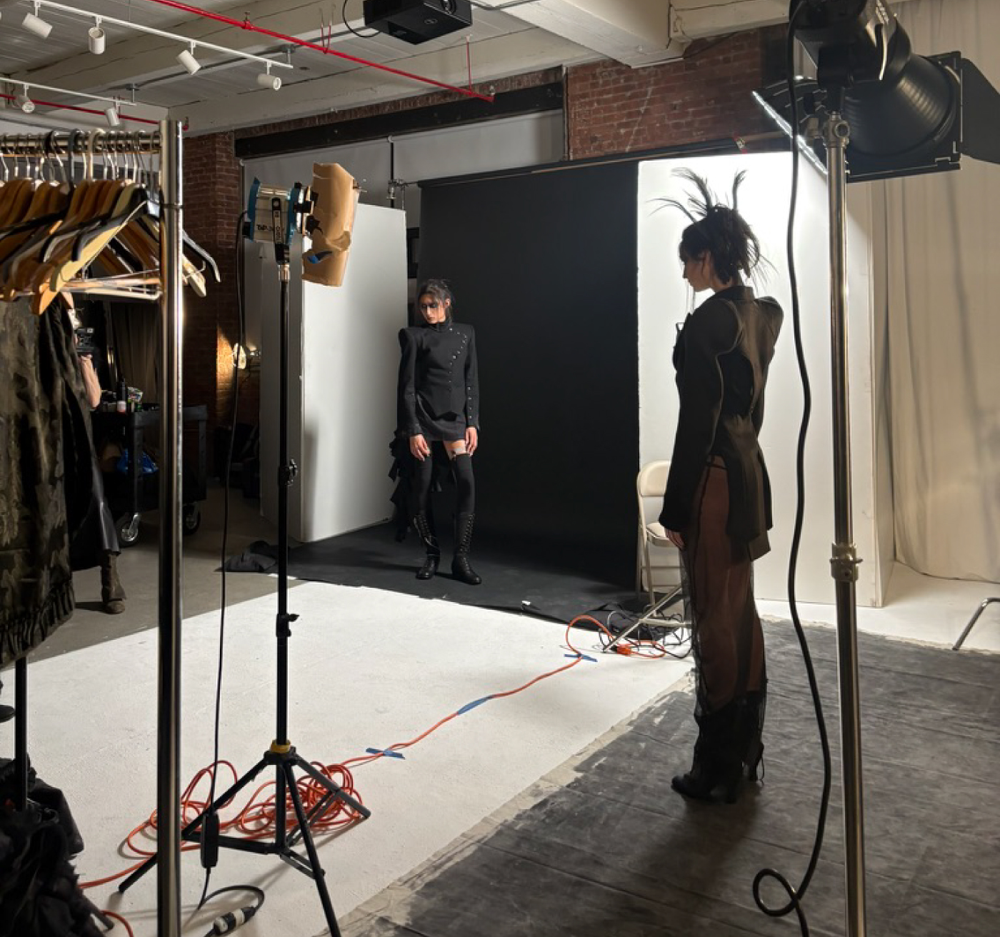 Fashion photo shoot in a studio, with a model posing against a backdrop while another model and lighting equipment are visible nearby.