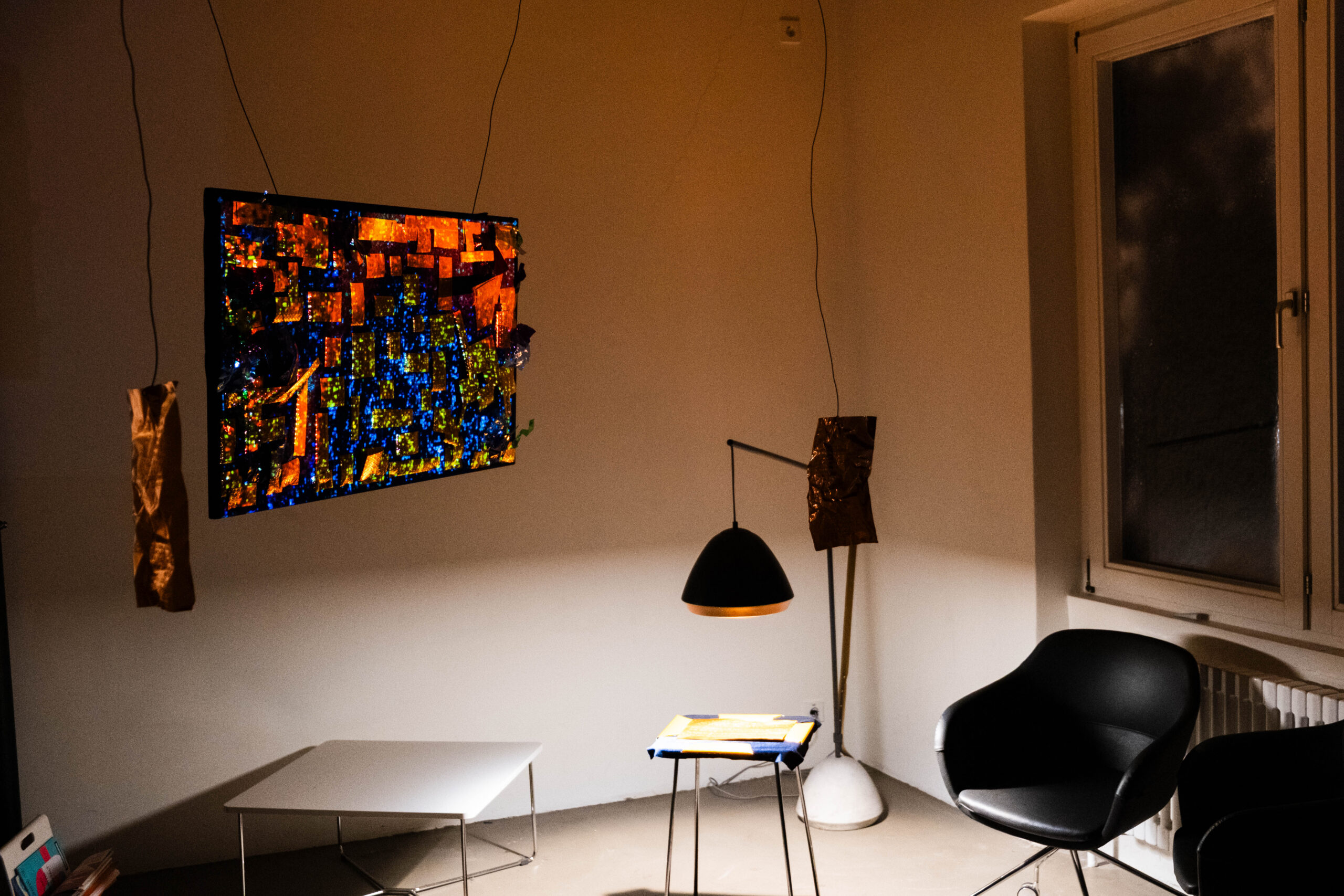 Student installation in a small room featuring a colorful illuminated wall piece, hanging materials, a chair, and a desk lit by one lamp.