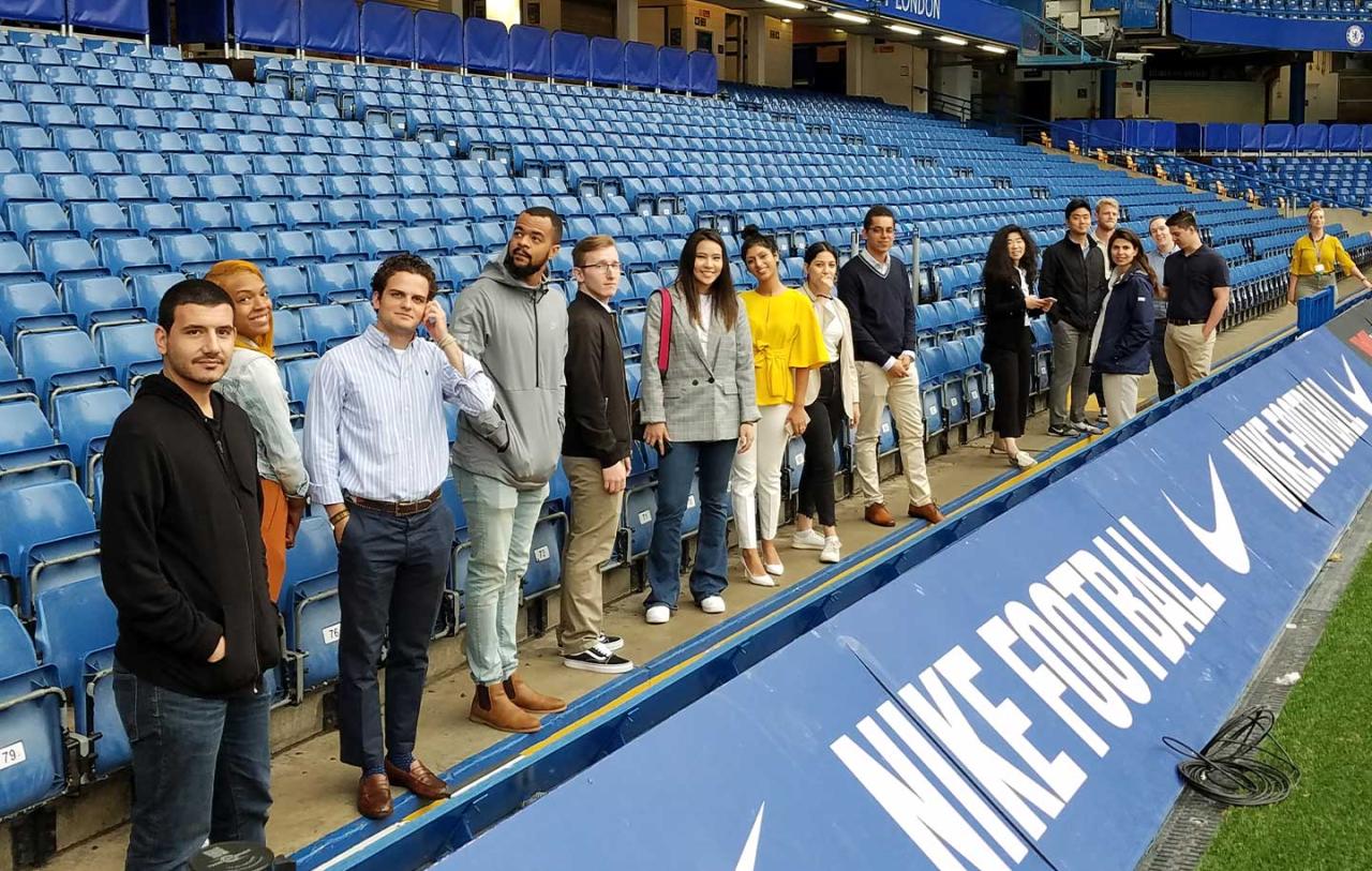 Group of sports management students standing in a line beside blue stadium seats, overlooking a field with Nike Football signage.