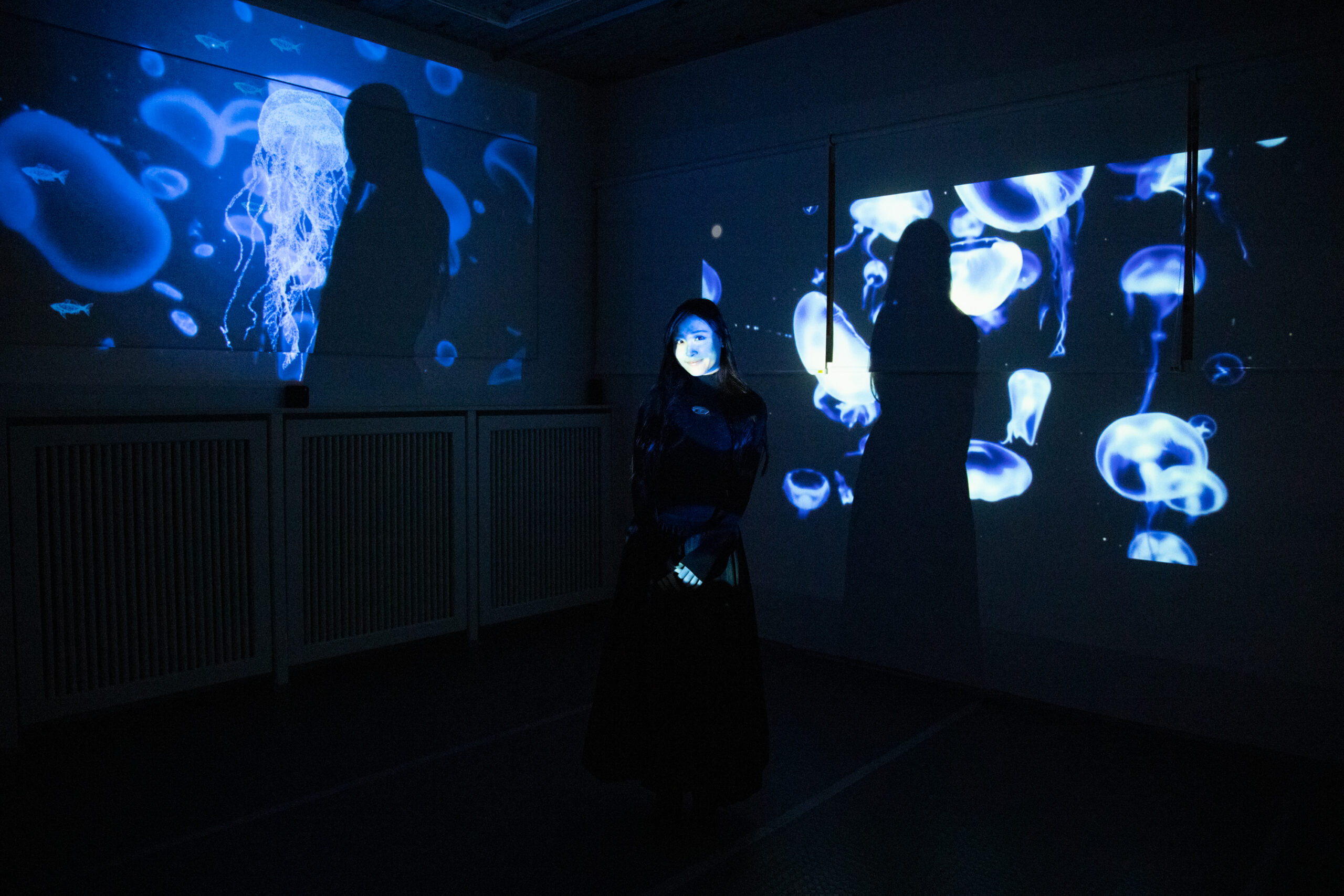 Student standing in a dark room with blue jellyfish projections on the walls, their face illuminated by the light.