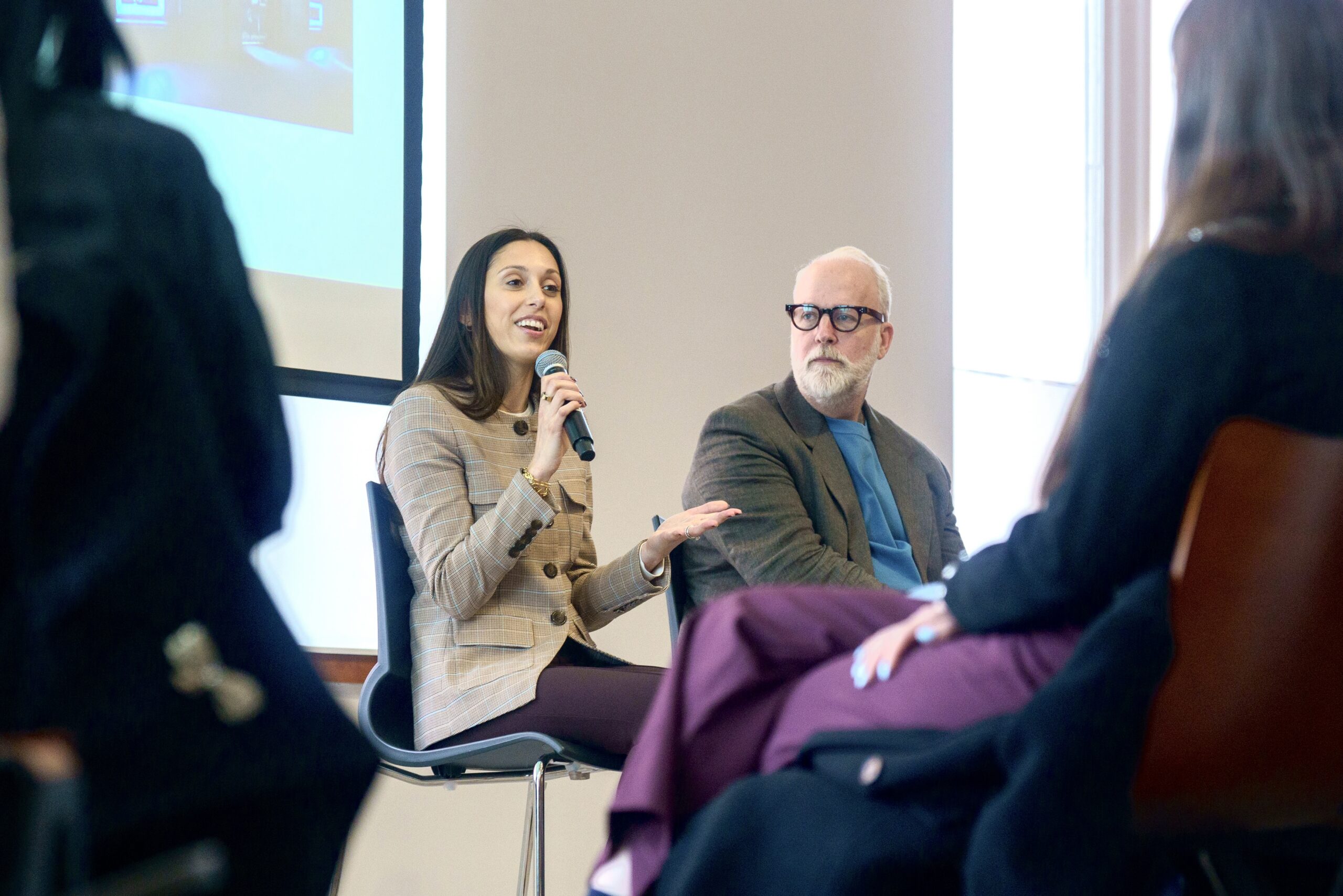 A guest speaks into a microphone during a panel discussion while another panelist listens, with audience members seated in the foreground.