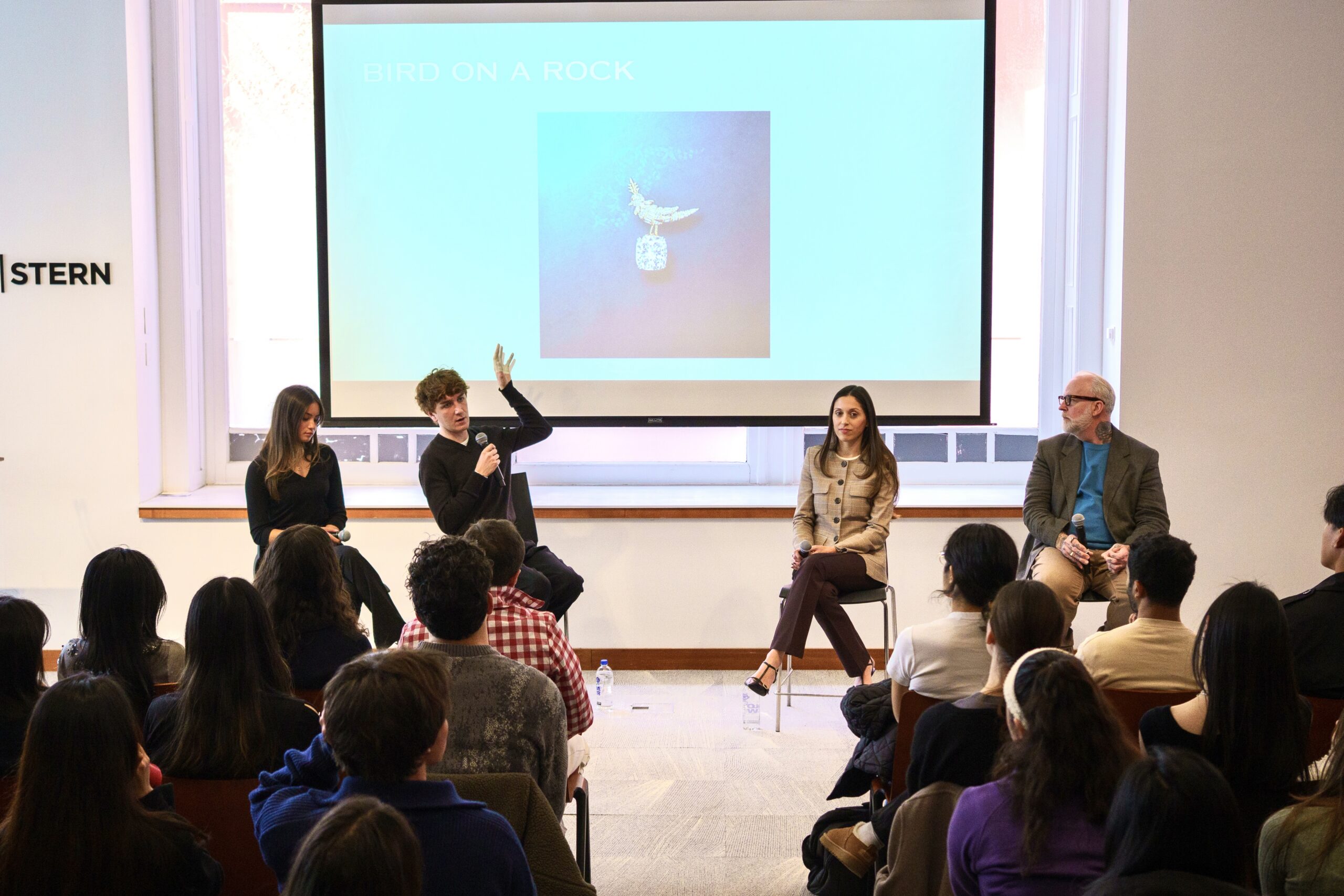 A panel of four speakers sitting and giving a presentation, while one speaker is addressing the crowd and a slideshow is displayed behind them.