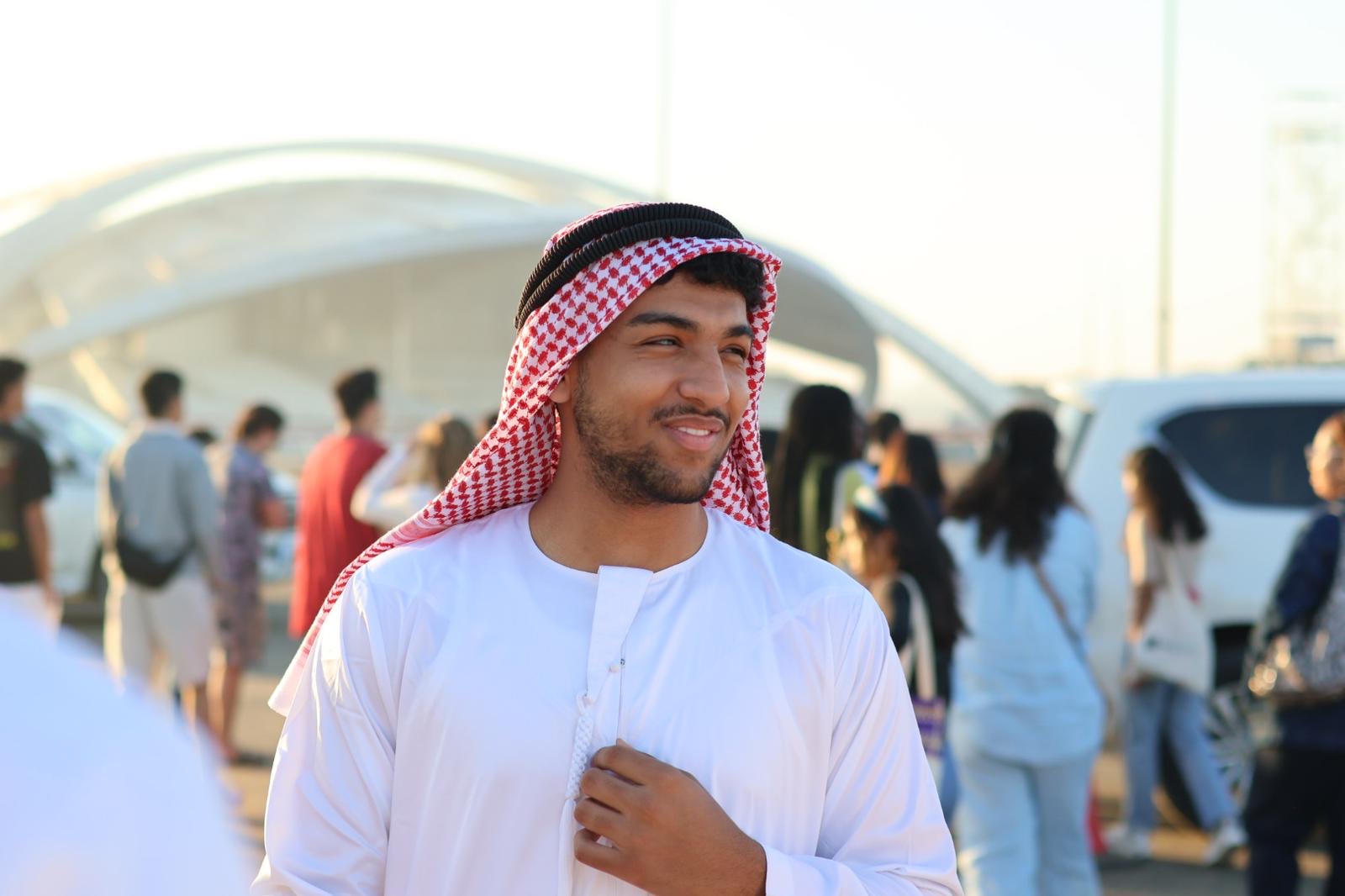 Saif, wearing a traditional robe and headscarf, stands outdoors in a crowd, with modern architecture blurred in the background.