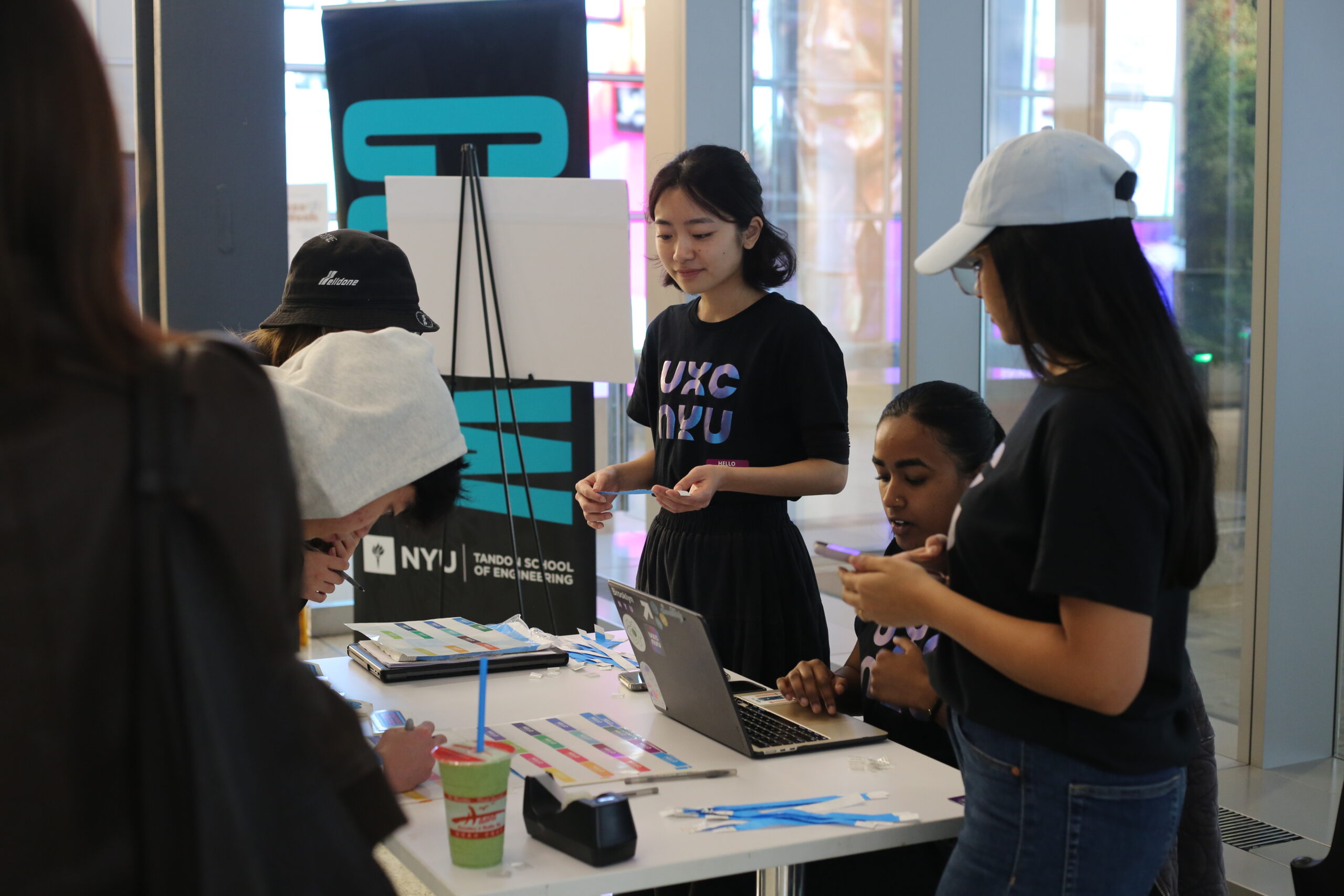 Students gather around a check-in table at an NYU event, using a laptop and name tags while volunteers assist attendees.
