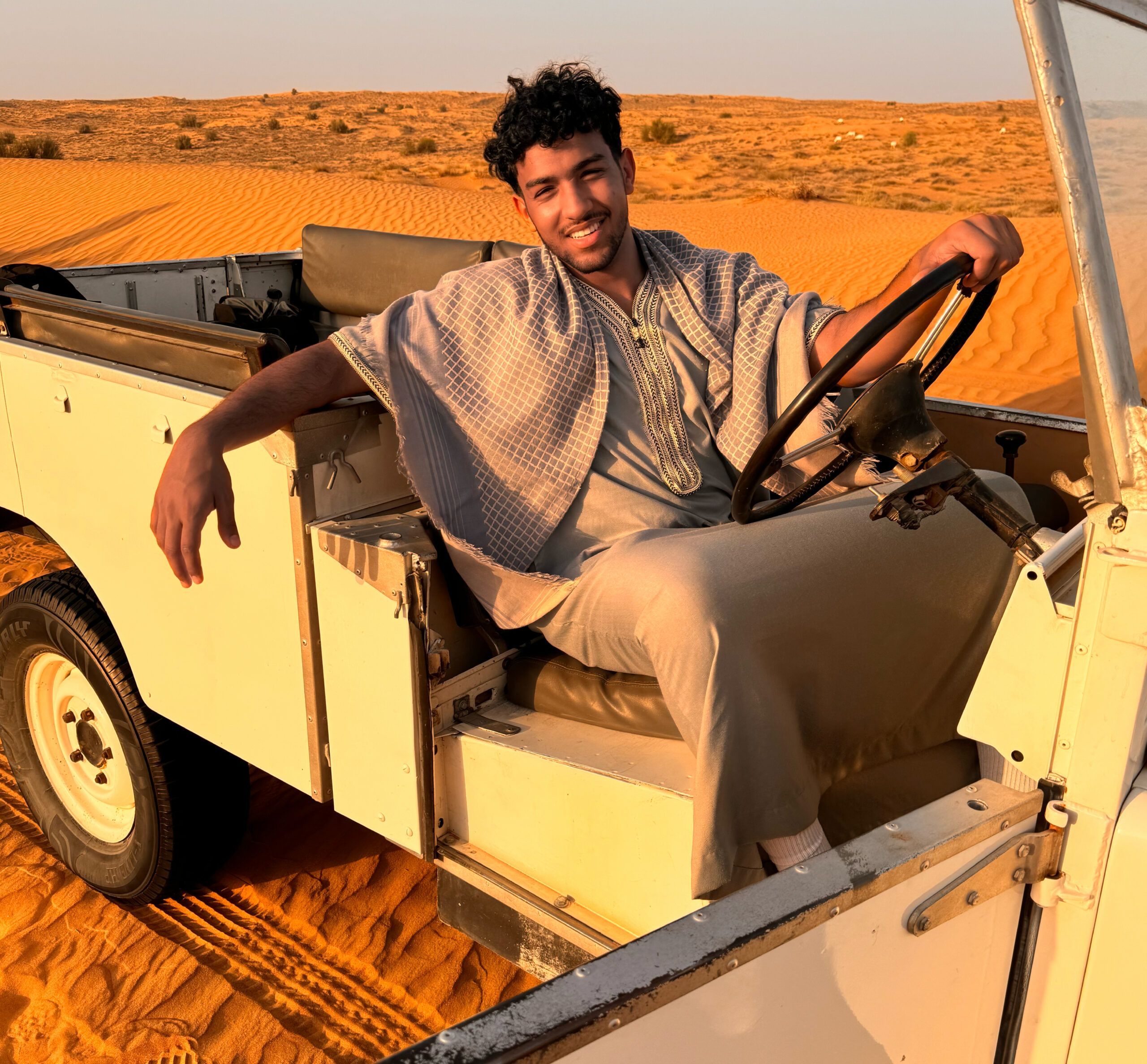 Saif, in traditional attire, sitting in an open-top vehicle on sand dunes during golden hour.