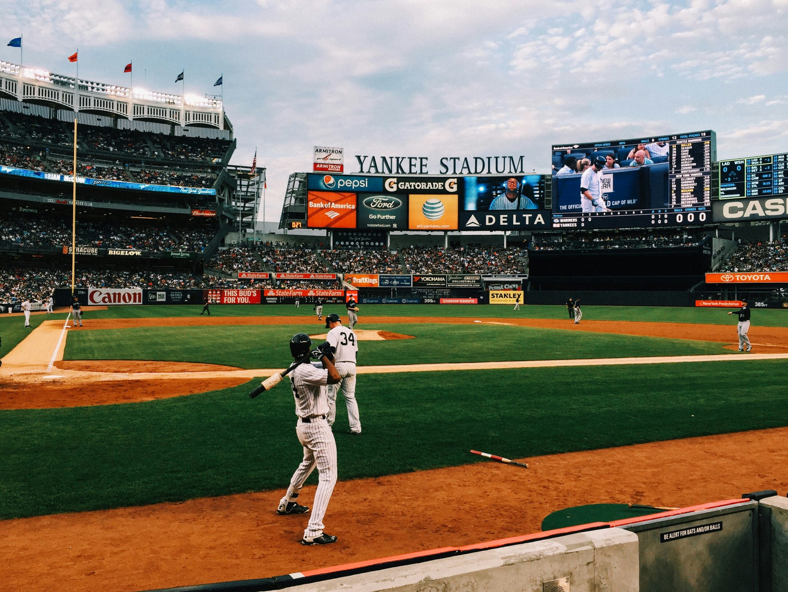 Baseball players warming up on the field at Yankee Stadium during a game, with fans in the stands and the scoreboard visible in the background.
