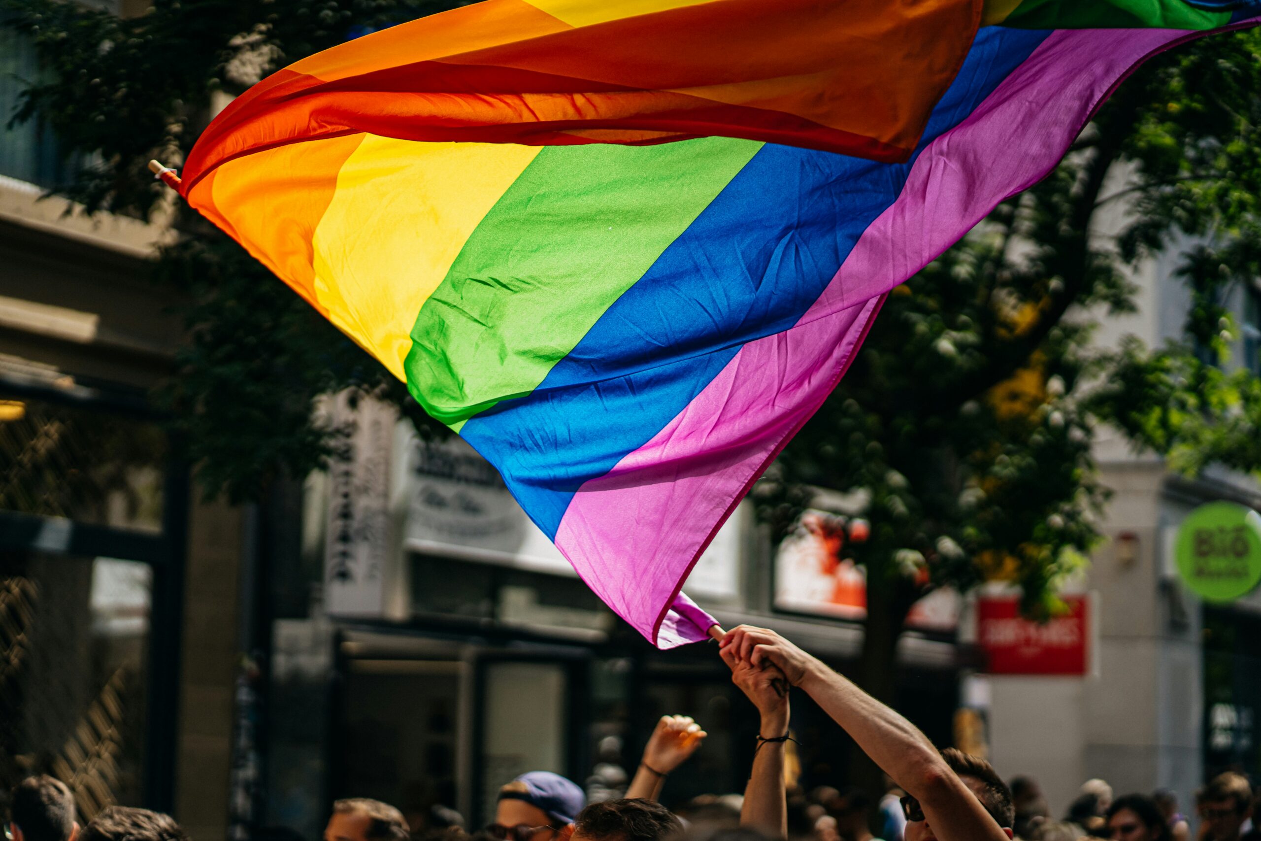 Someone waves a large rainbow flag through the air on a city street.