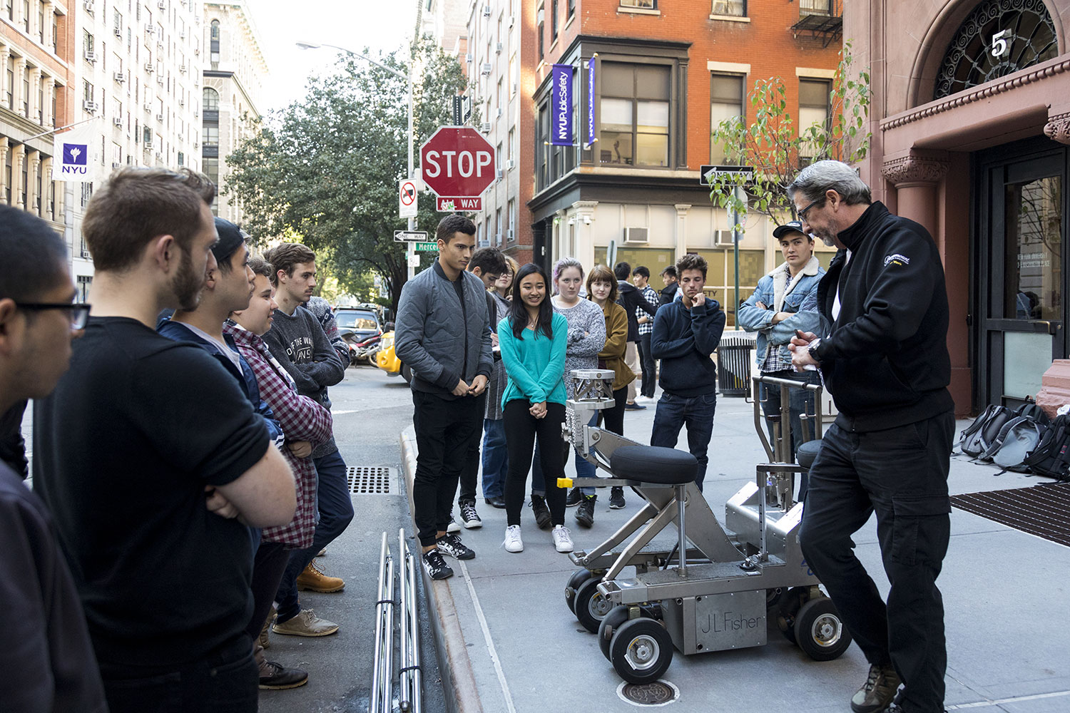 A group of students gathers on a sidewalk as an instructor demonstrates professional film production equipment near NYU campus buildings.