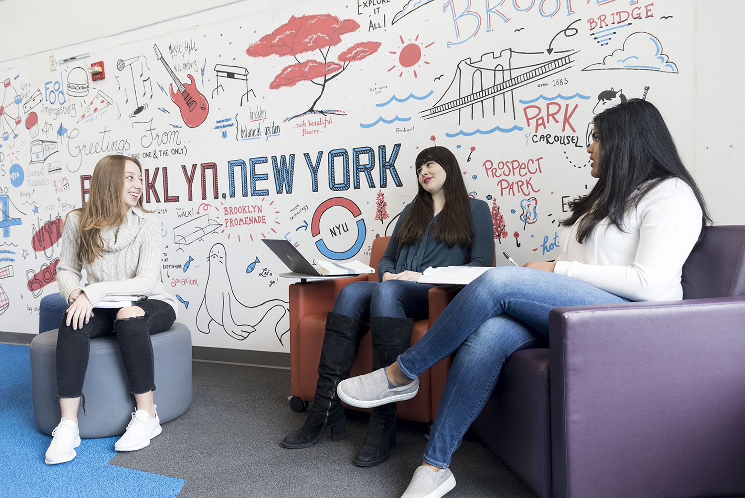 Three students sit together in a lounge area, collaborating in front of a colorful mural featuring landmarks inspired by New York City.