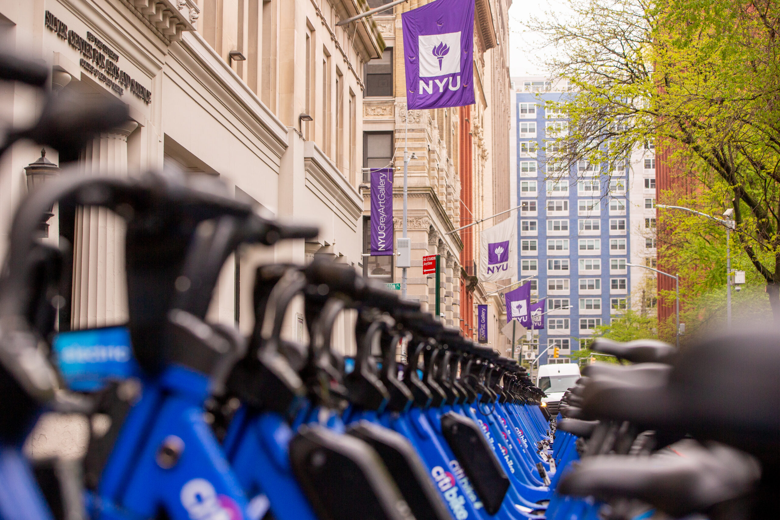 A row of blue Citi Bikes lines a city street near NYU campus buildings, with purple NYU banners hanging overhead.