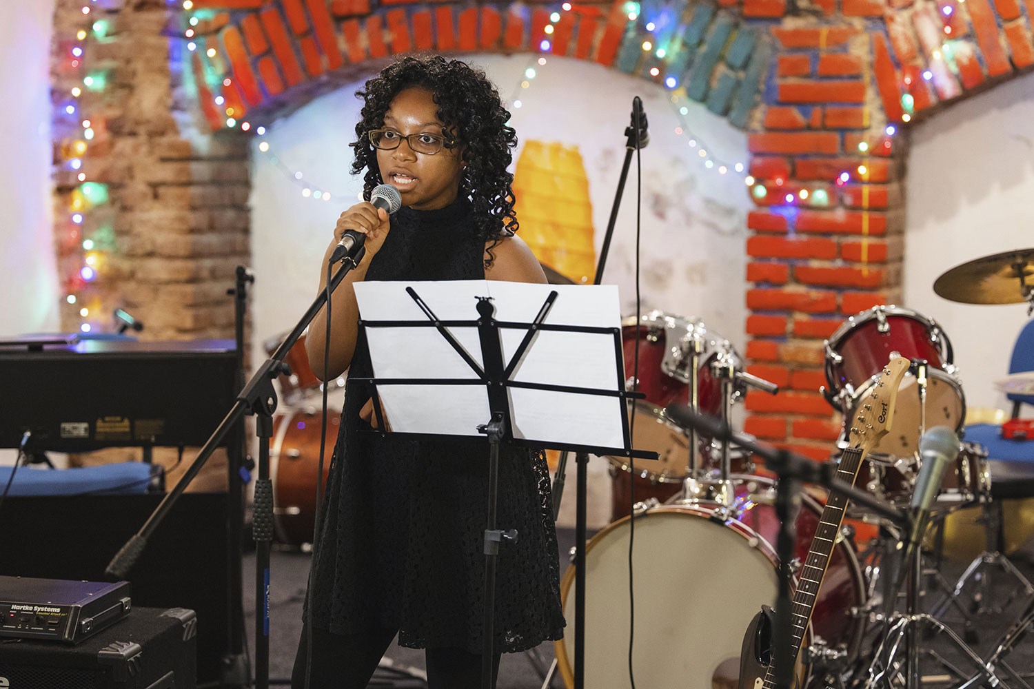 A student speaks into a microphone in front of musical instruments and colorful string lights in an intimate performance space.