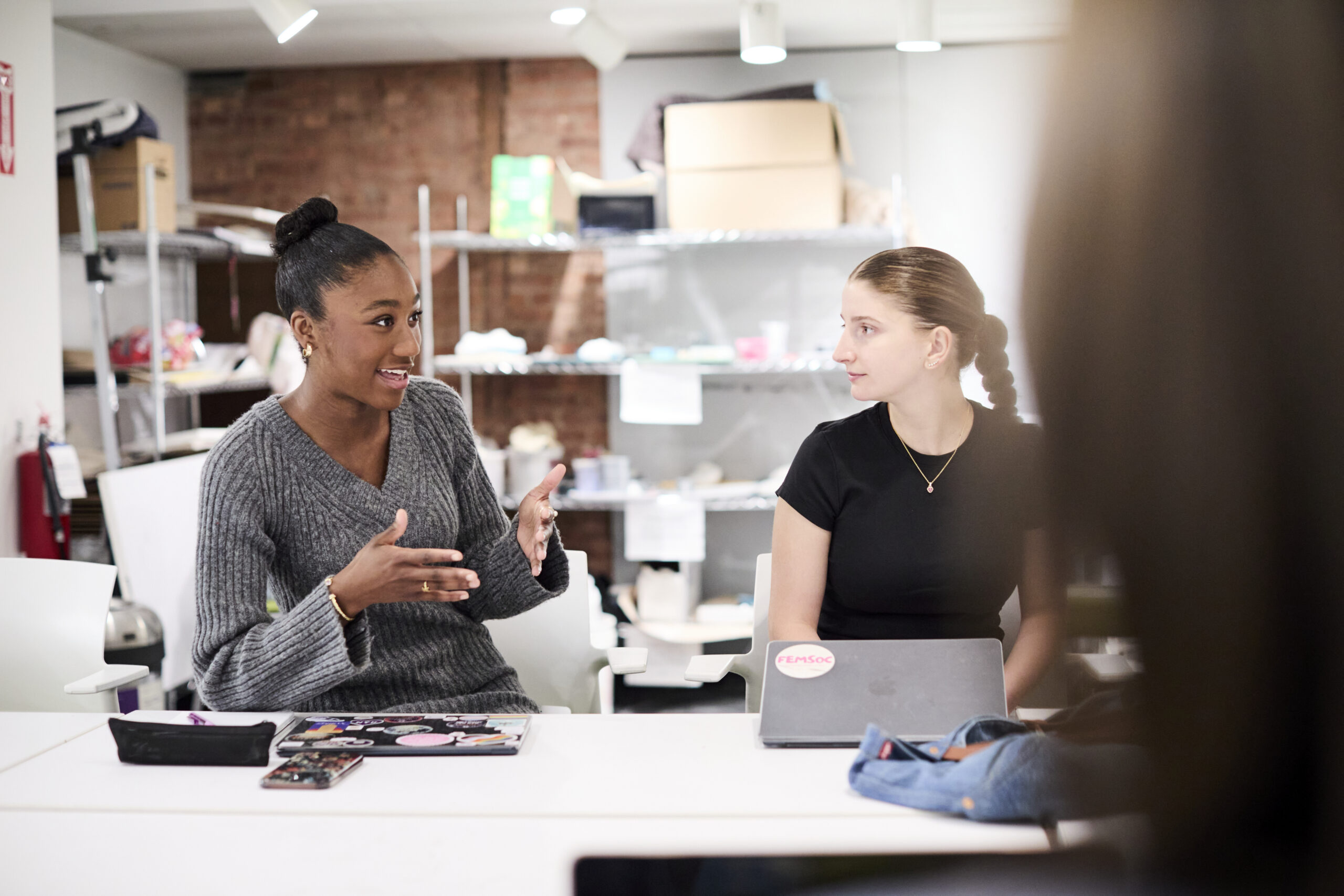Two students sit at a studio table, engaged in discussion.