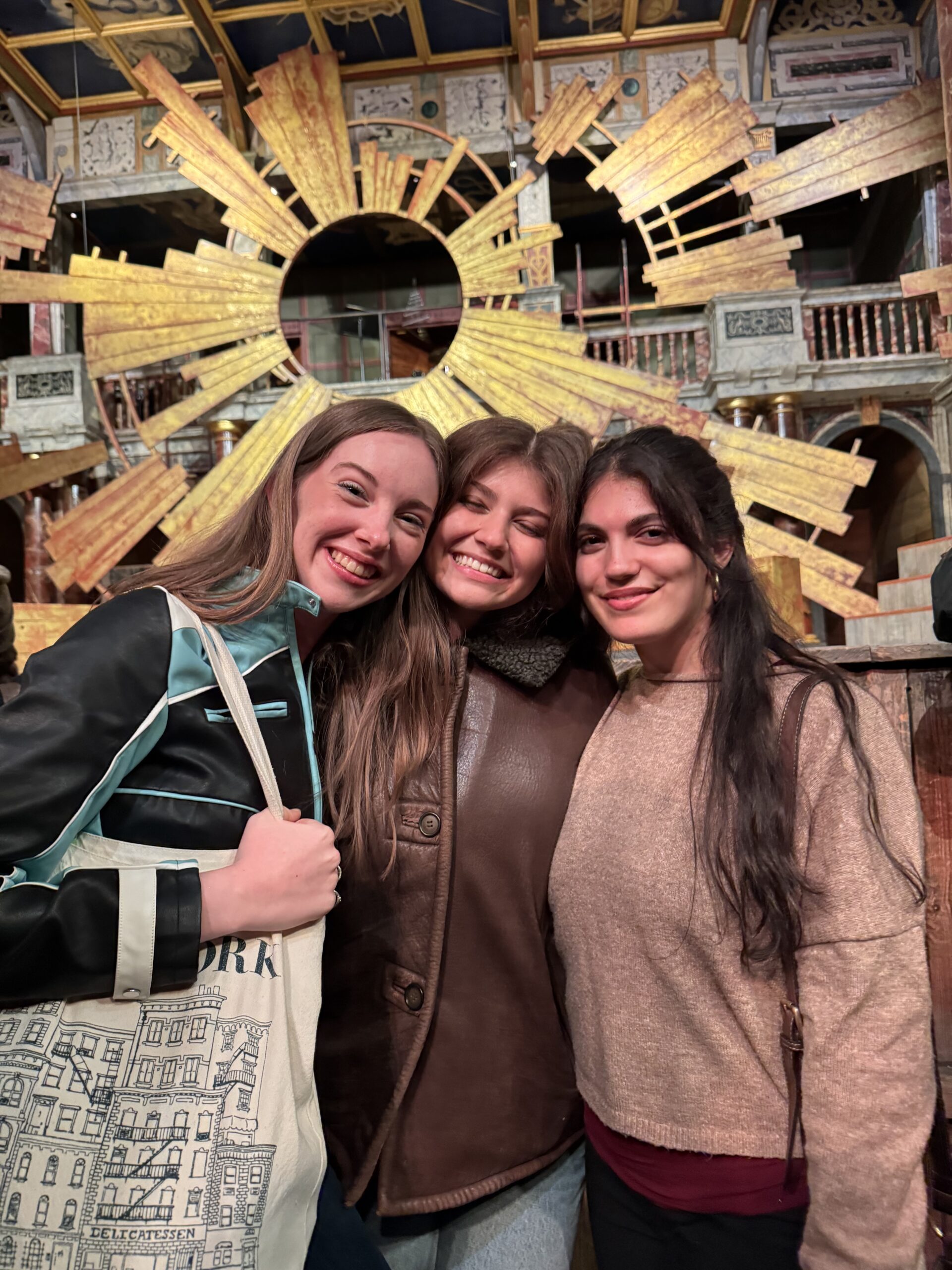 Three students hug and smile in front of a stage.