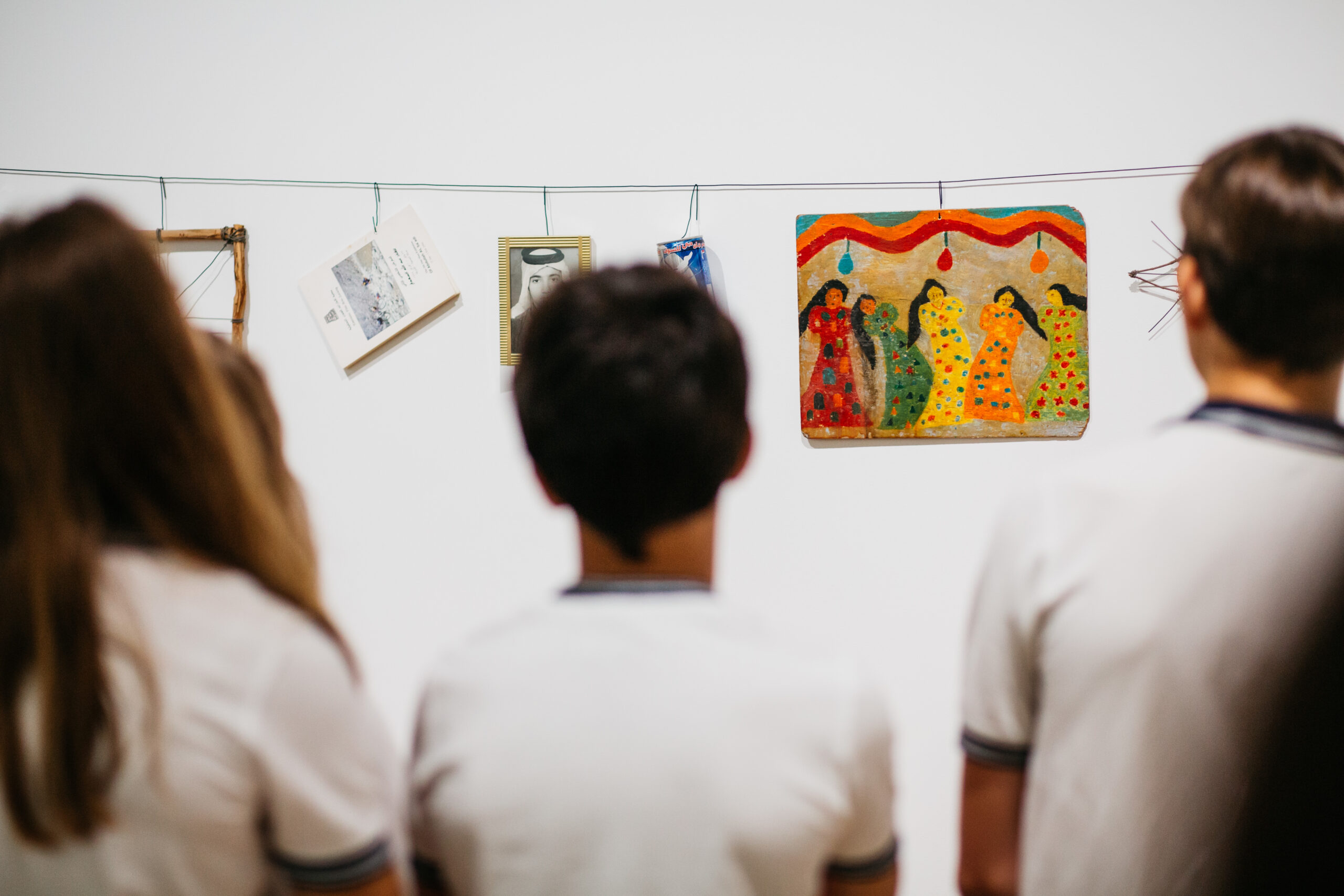 Students view and discuss artwork displayed in a gallery setting featuring colorful paintings, photographs, and mixed-media pieces.