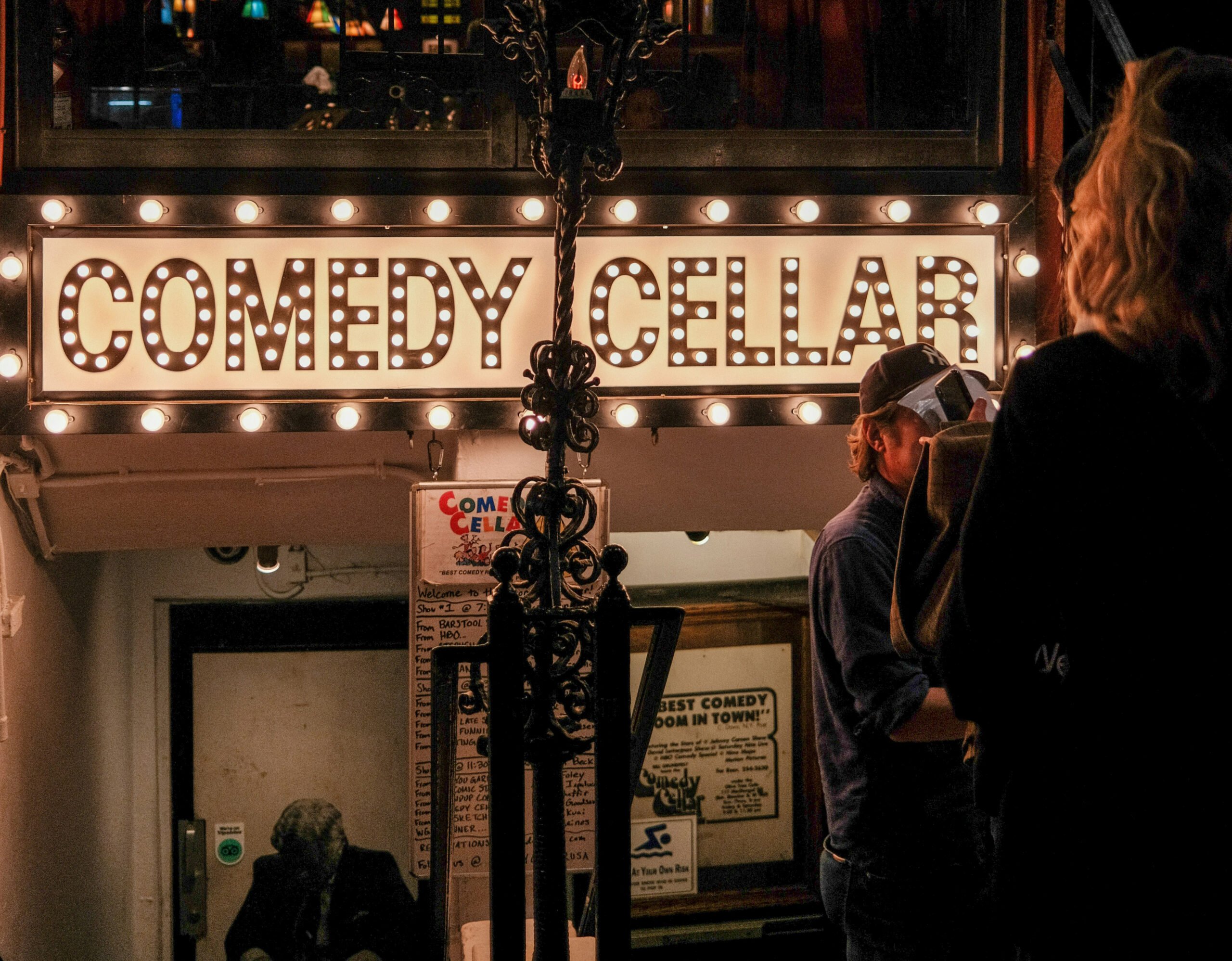 The illuminated entrance to the Comedy Cellar glows with marquee lights as people gather outside the iconic New York City comedy club.