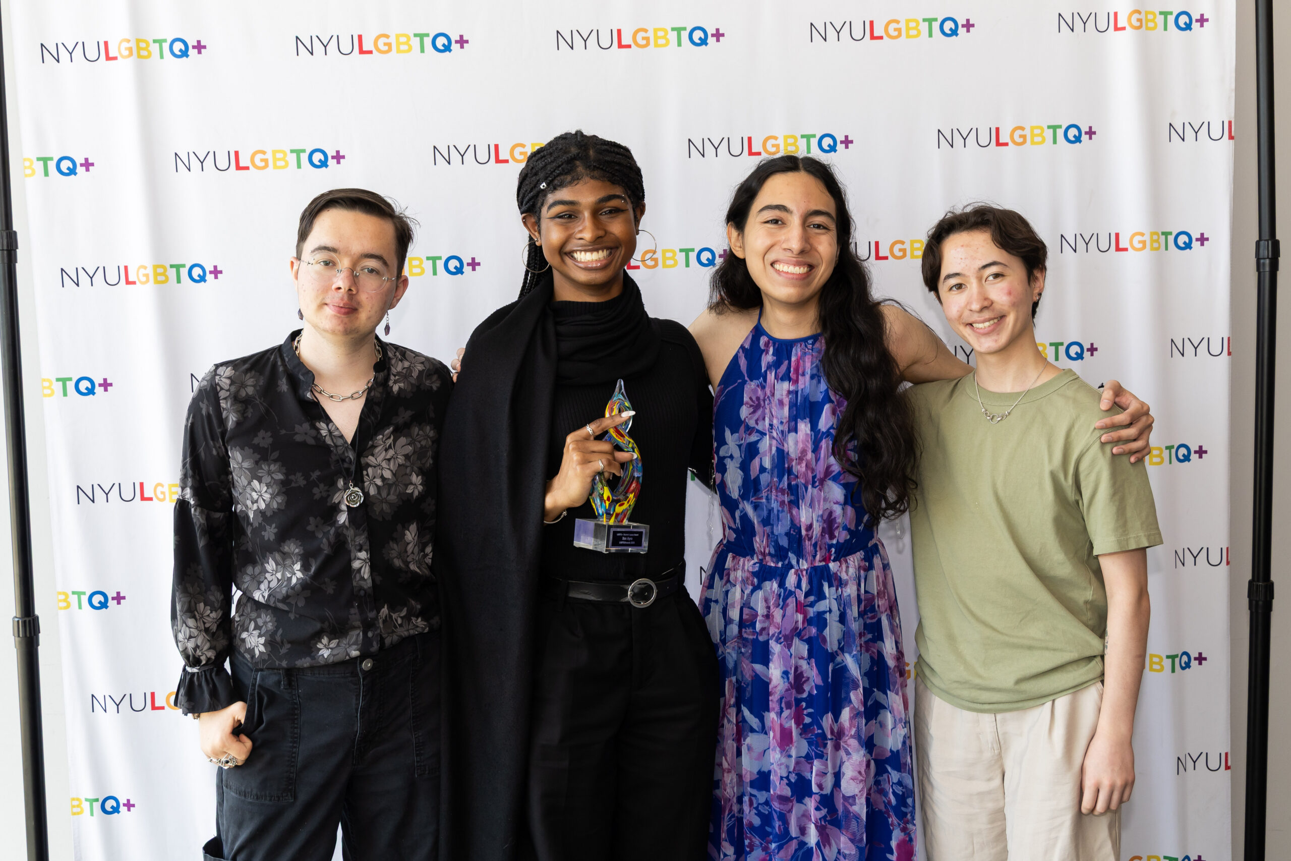 Four smiling students stand together in front of an NYU LGBTQ+ backdrop.