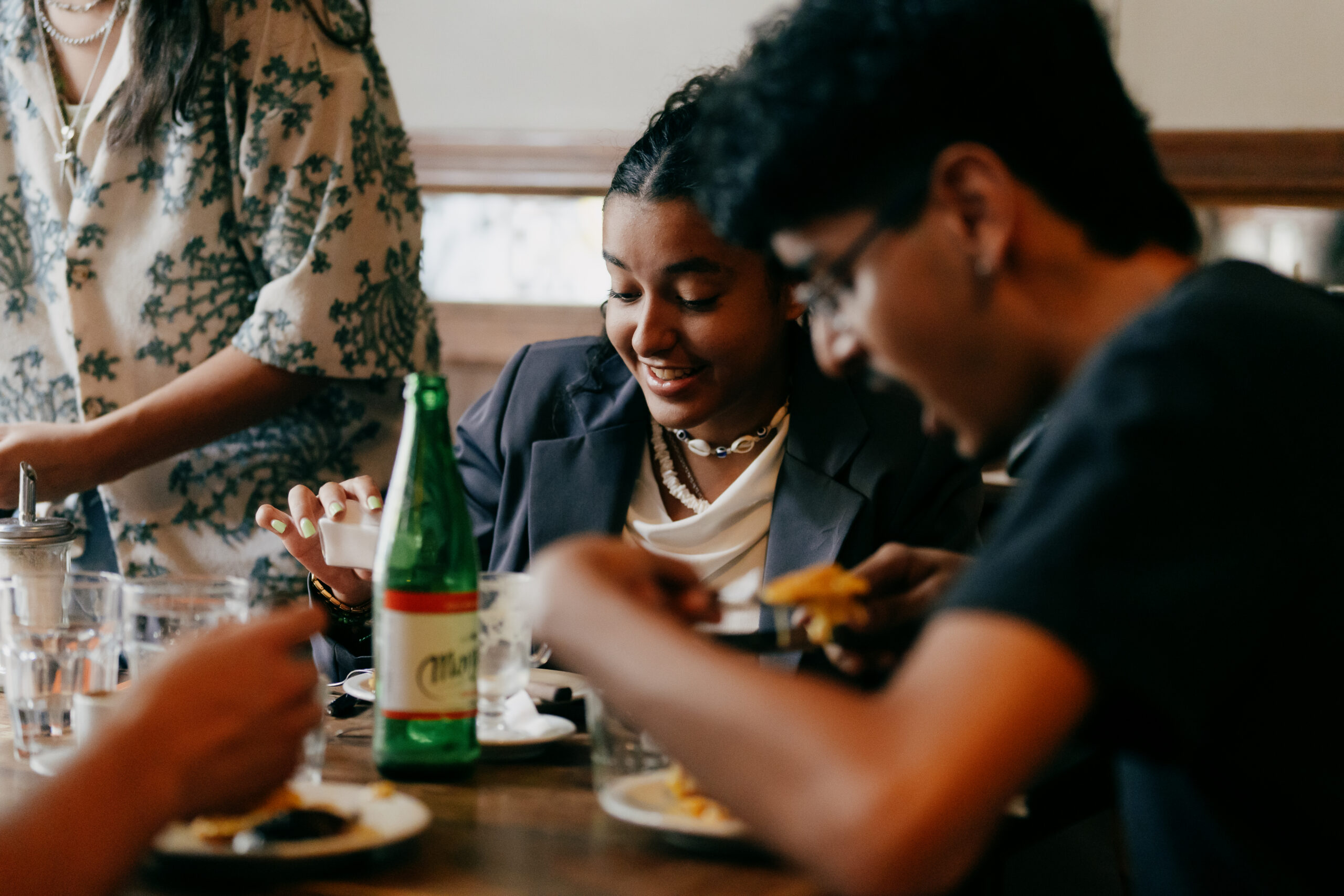 Students share a meal around a table, smiling and talking in a casual restaurant setting.