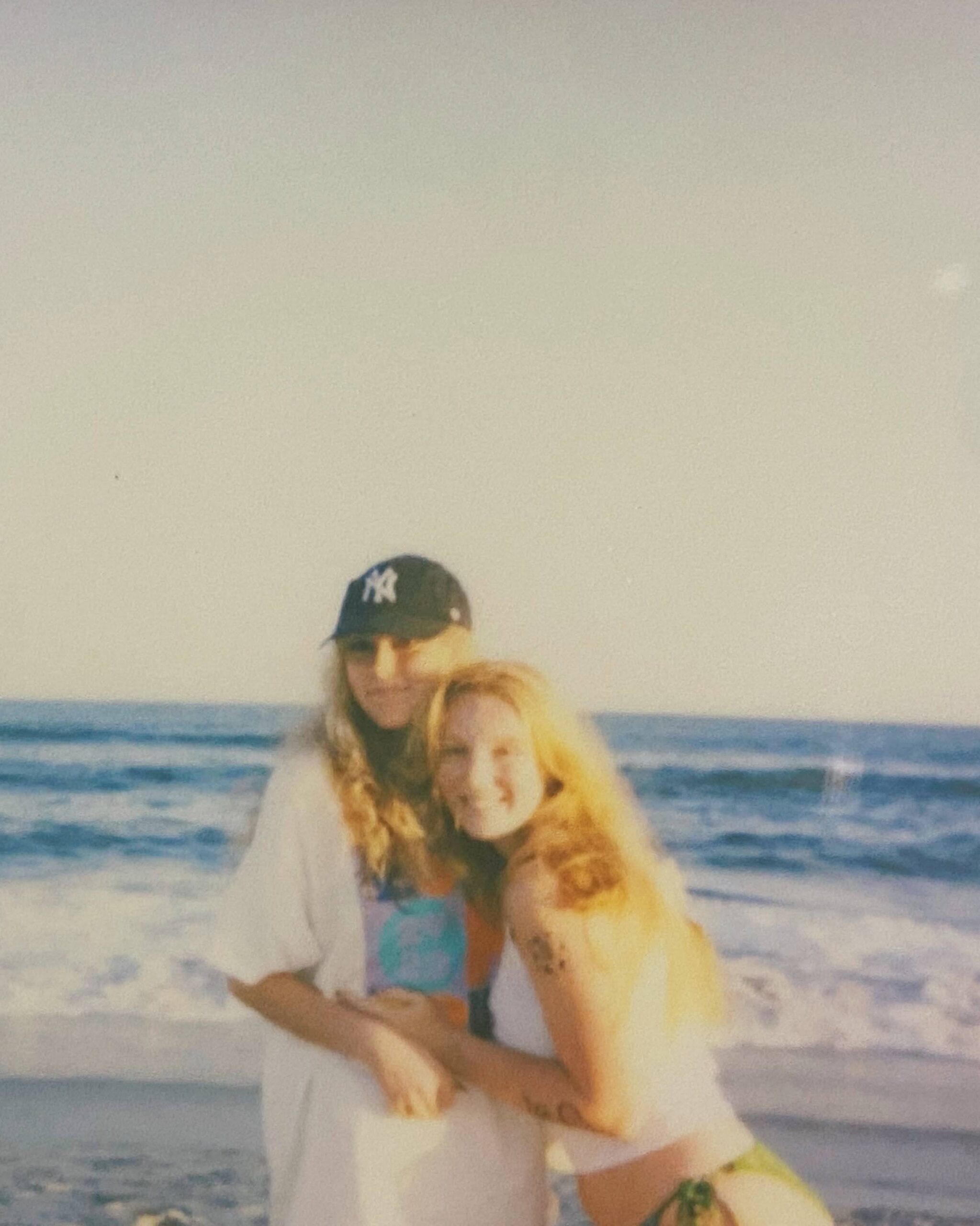 Two girls wearing casual clothes pose happily on a beach with big waves during sunset.