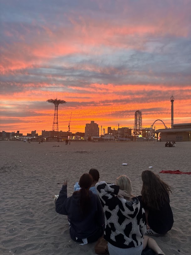 Four friends sitting on the Coney Island beach and watching an orange & pink sunset over the amusement park.