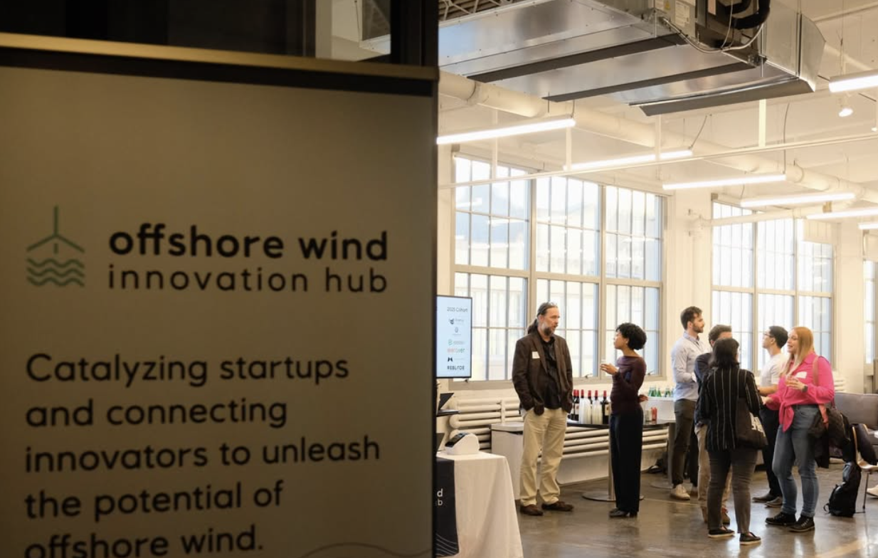 Attendees gather and converse in a bright event space with large windows, behind a sign for the Offshore Wind Innovation Hub.