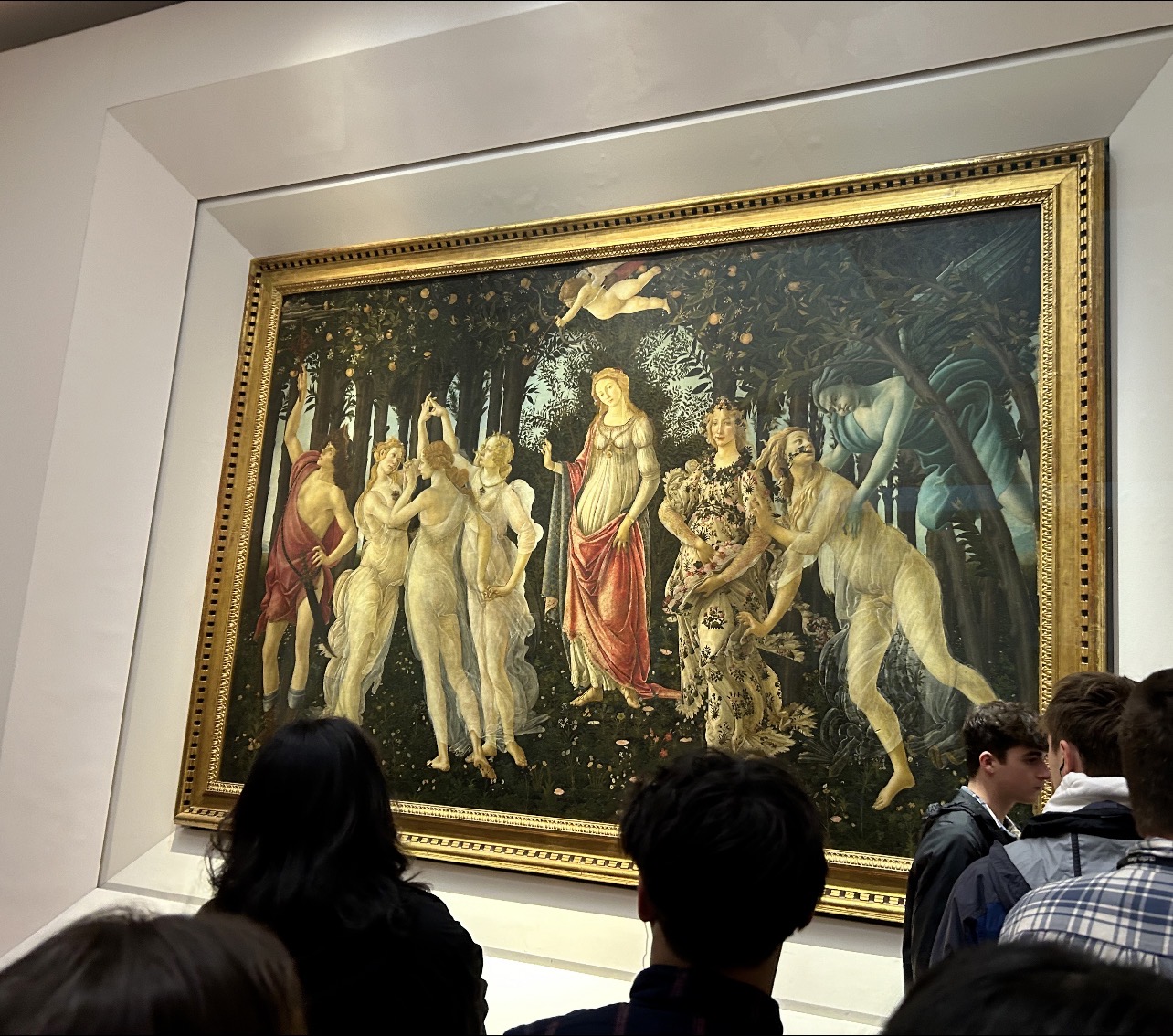 an early renaissance painting depicting nine mythological gigures in an orange forest, a blue-faced God on the right holding a woman, three women dancing with linked hands on the left, a blindfolded cupid hovering above, a women in the middle of the painting looking at the audience.