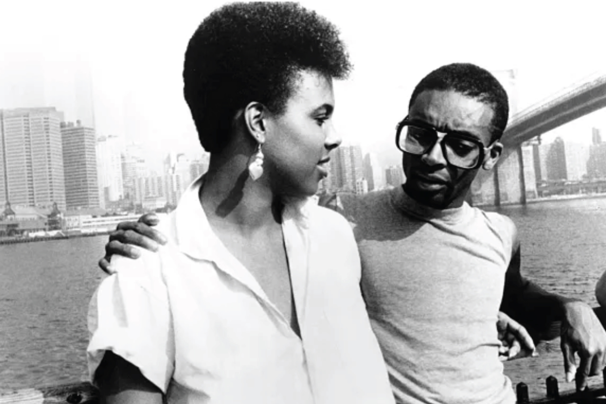 Black-and-white still from She's Gotta Have It showing a woman and a man seated by the waterfront, facing each other in conversation, with the Manhattan skyline and Brooklyn Bridge in the background.