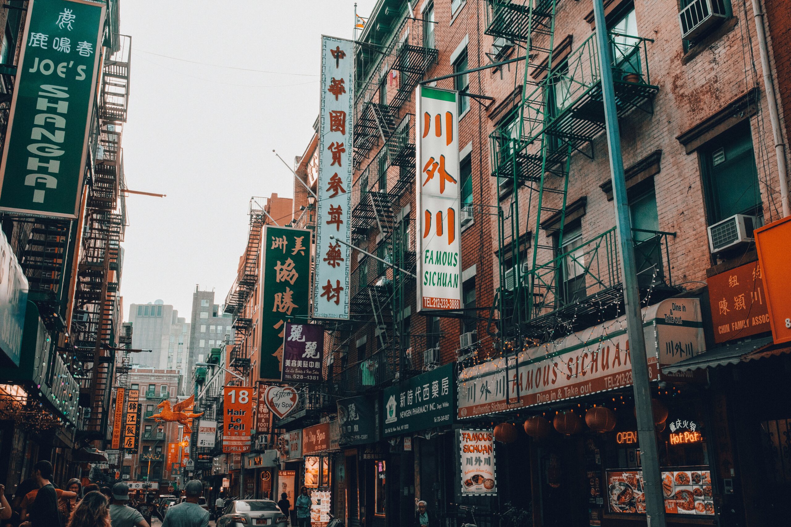 A busy street in New York City's Chinatown with pedestrians walking along the sidewalks beneath a dense collection of colorful storefront signs and awnings.