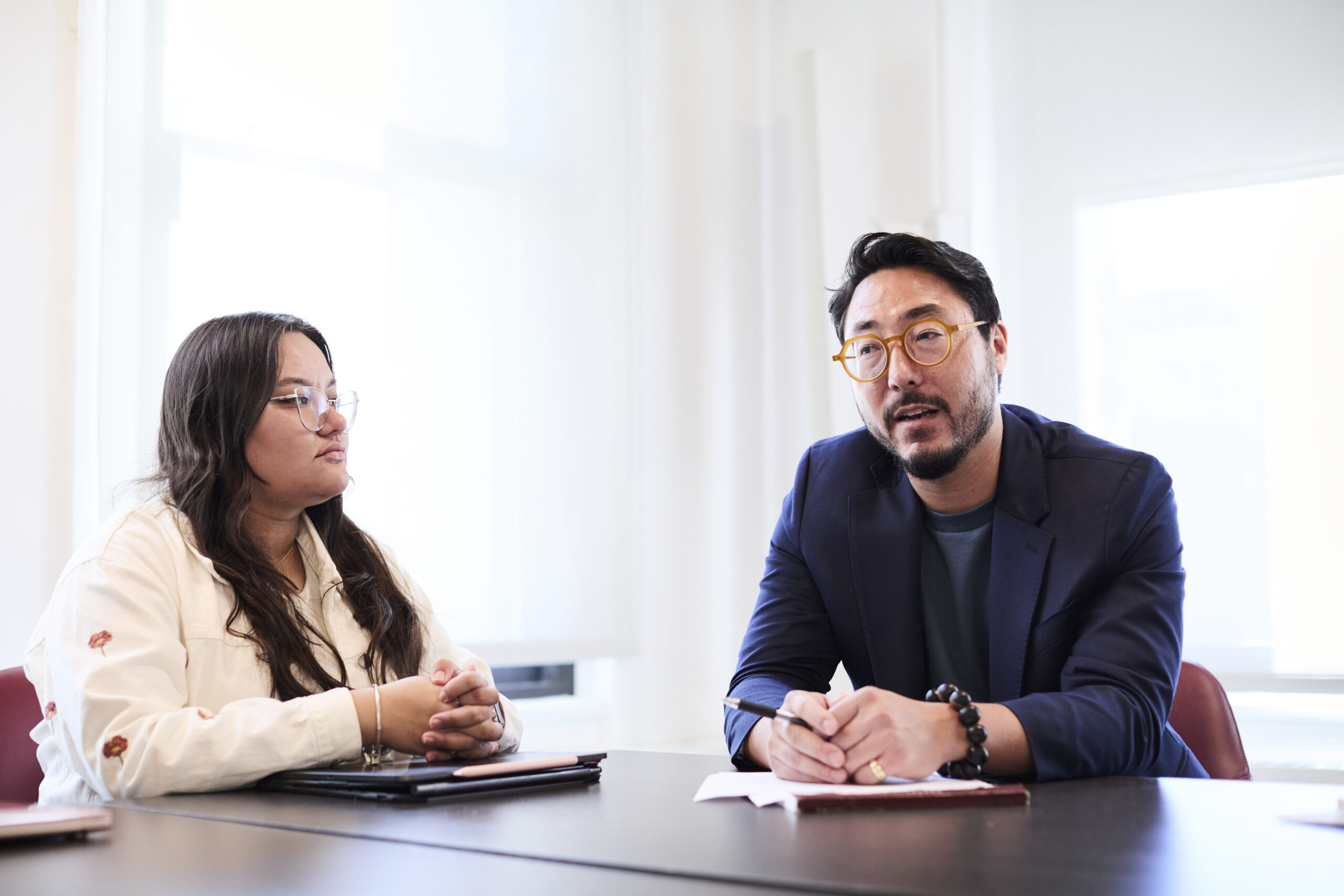 Both seated at a conference table, a student listening intently to a professor.